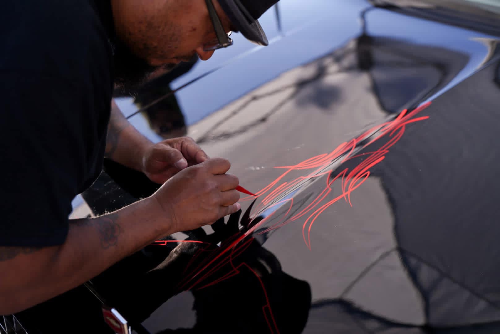 Person drawing red lines on a shiny black surface, possibly a car, with a reflection of a pole and sky. Word count: 20