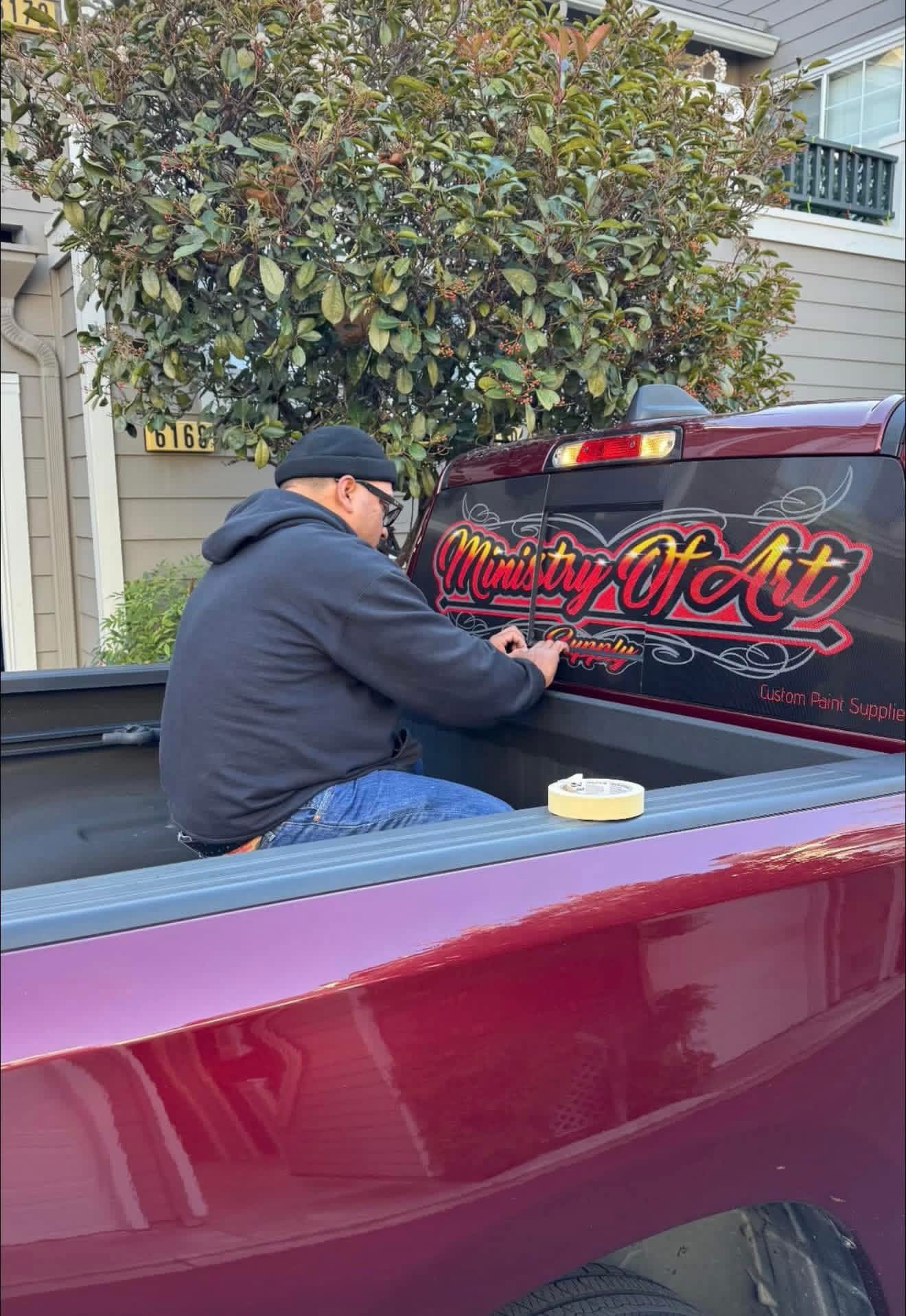Person sitting in truck bed, working on a vehicle with a tree and house in background.