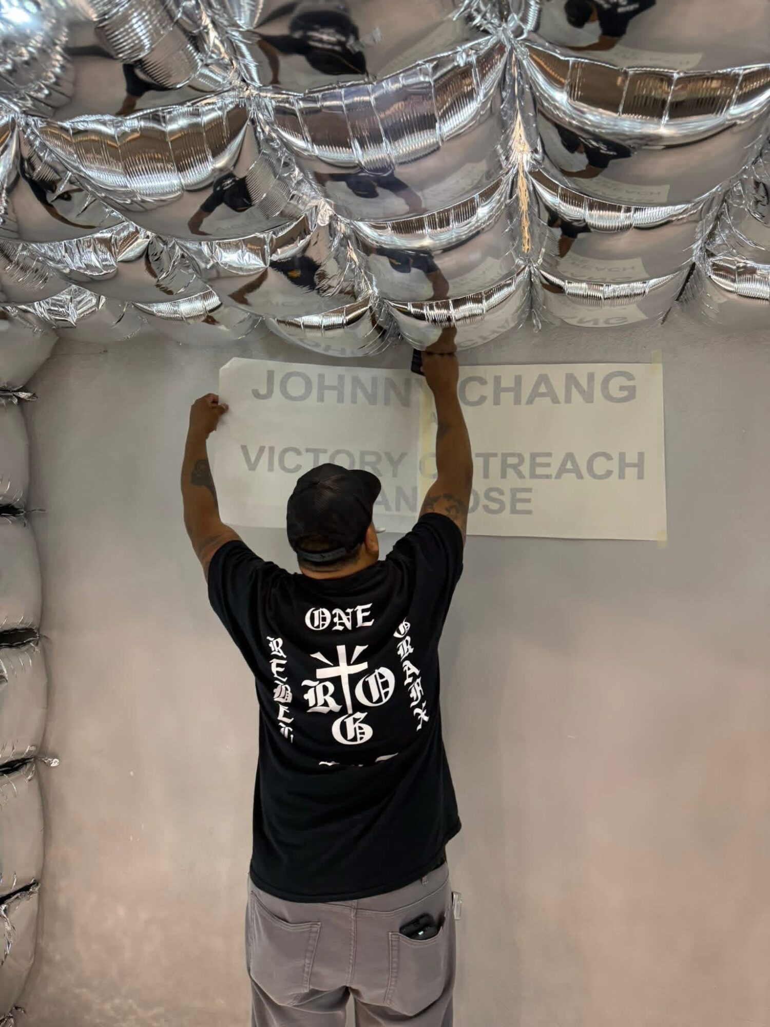 Person hanging a sign on a wall with silver balloons overhead, wearing a black T-shirt and gray pants.