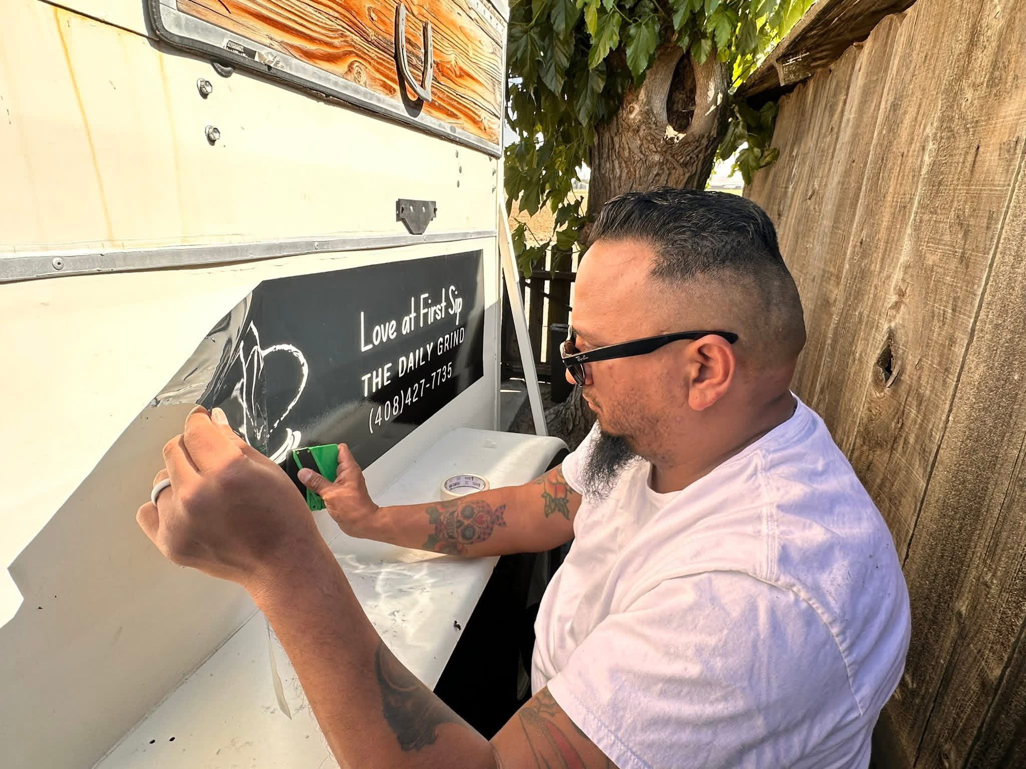 Man with glasses and tattoos in a white shirt writing on a white surface outdoors, with a wooden fence and tree in background.