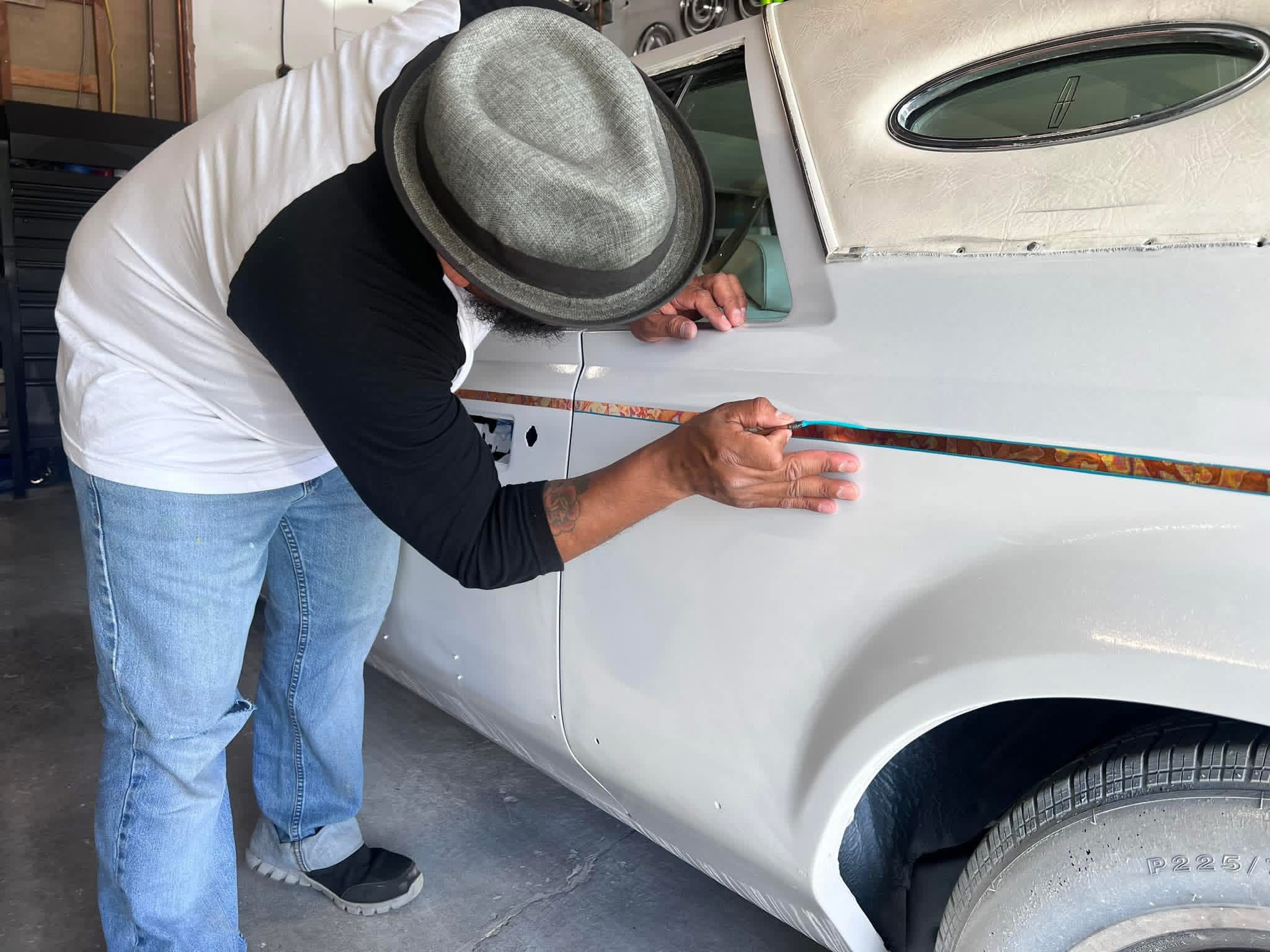 Person measuring car door with a ruler, wearing a hat and casual clothing, in a garage or workshop setting.