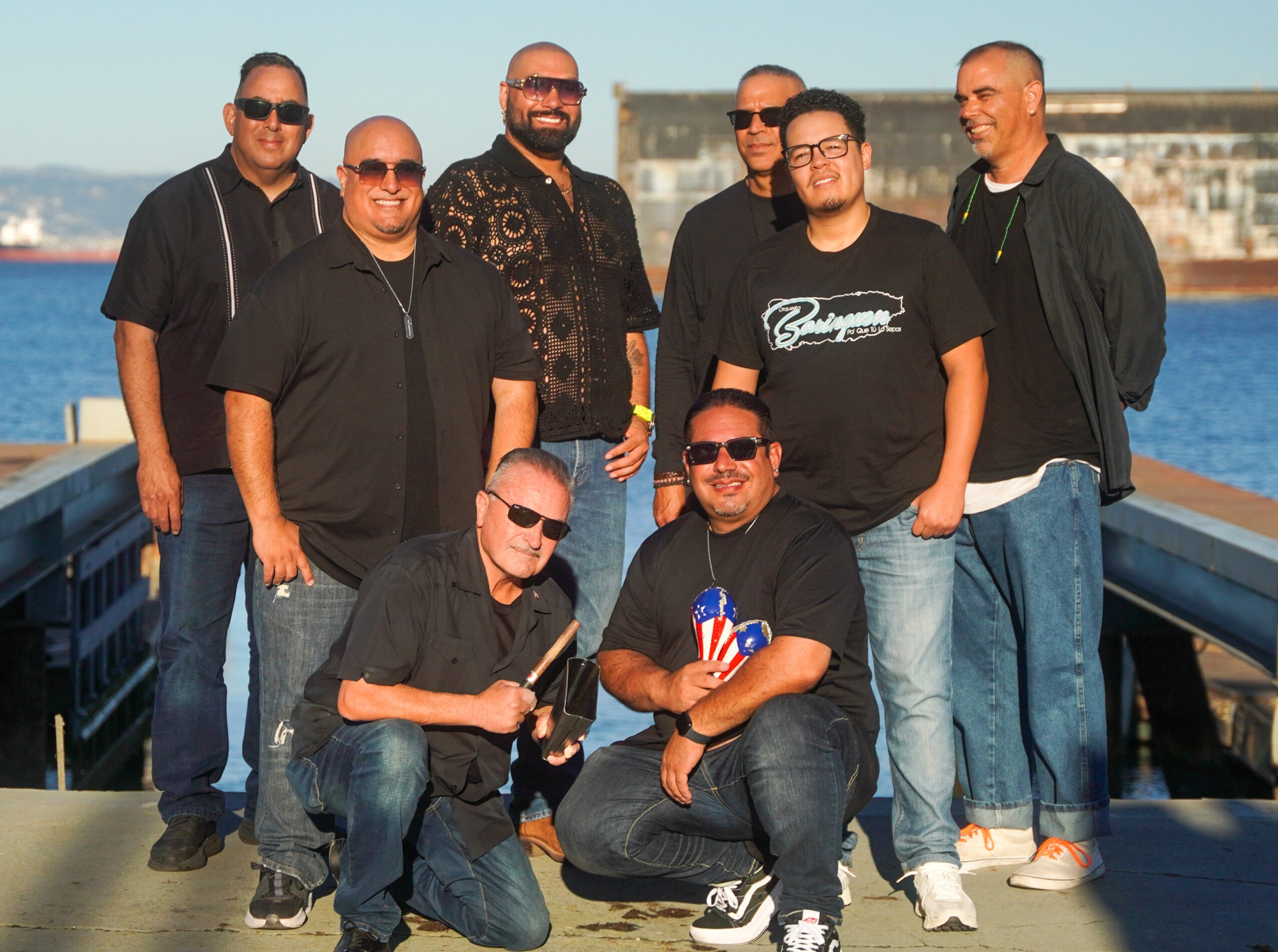 Group of nine people standing outdoors near water, some wearing sunglasses, smiling, with a building in the background.