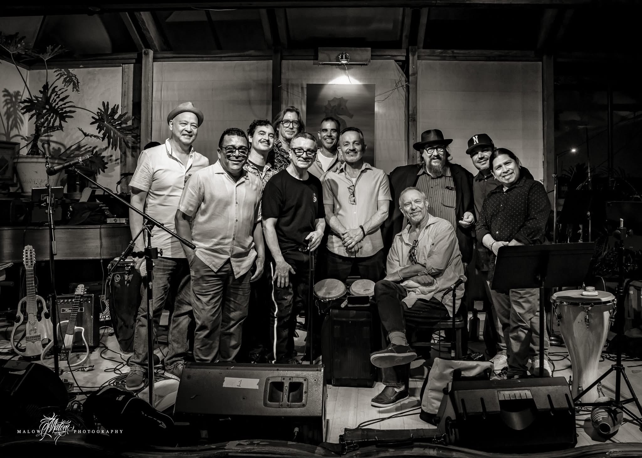 Group of ten people posing indoors with musical instruments and equipment, some smiling, in a casual setting.