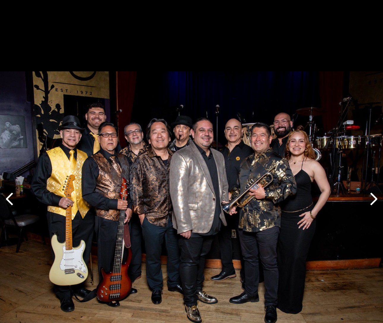 Group of ten musicians on stage, some holding instruments, in a dimly lit venue with a dark background.