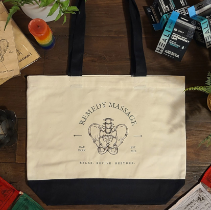 Cream-colored tote bag with black handles and a logo reading 'Remedy Massage' on a wooden surface.
