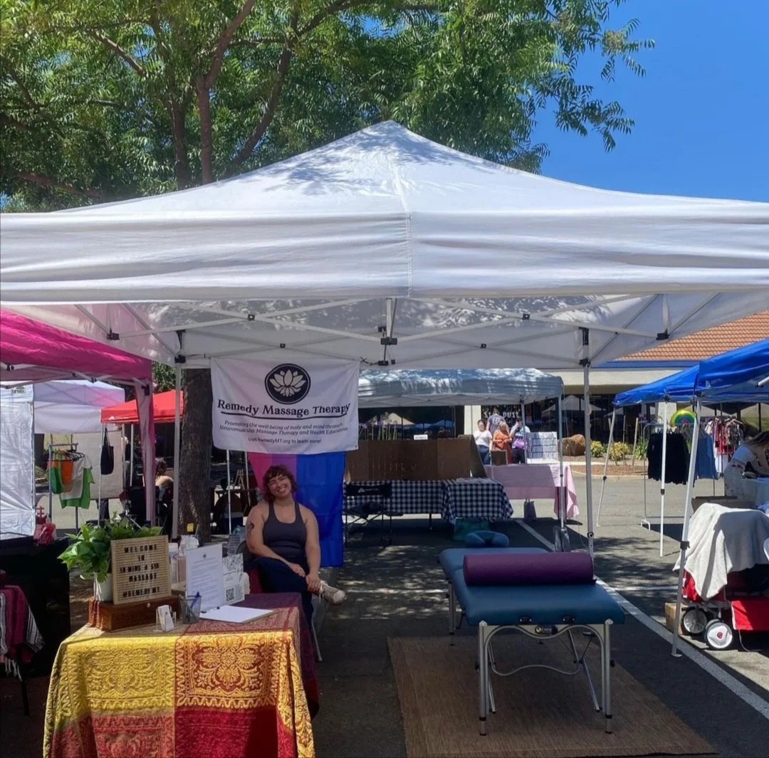 Woman sitting at a booth under a white canopy at an outdoor market, with tables and other tents around.
