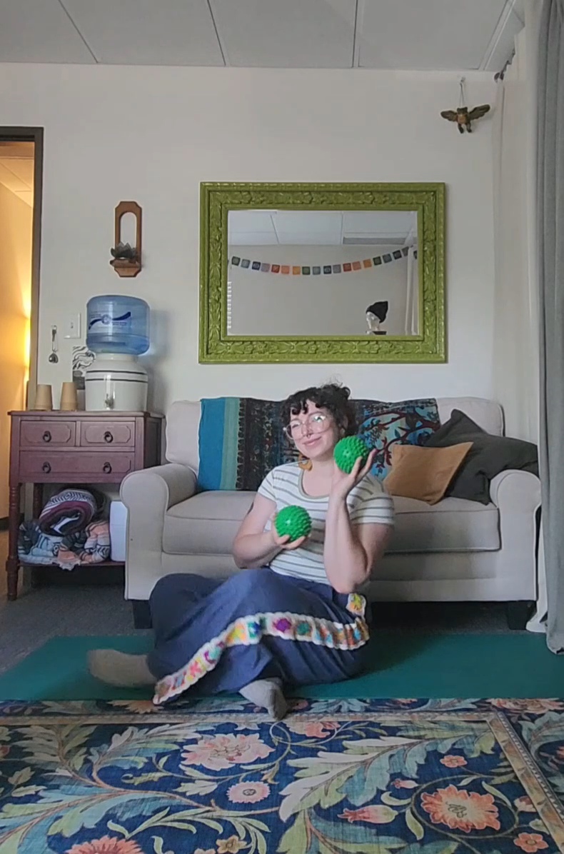 Person sitting on floor holding green maracas in living room with sofa, mirror, and water dispenser, colorful decor, and patterned rug