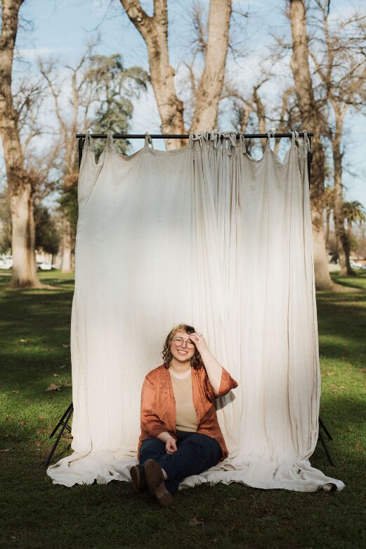 Person sitting on grass in front of a white curtain backdrop outdoors, trees in background, smiling, wearing glasses and casual clothing.
