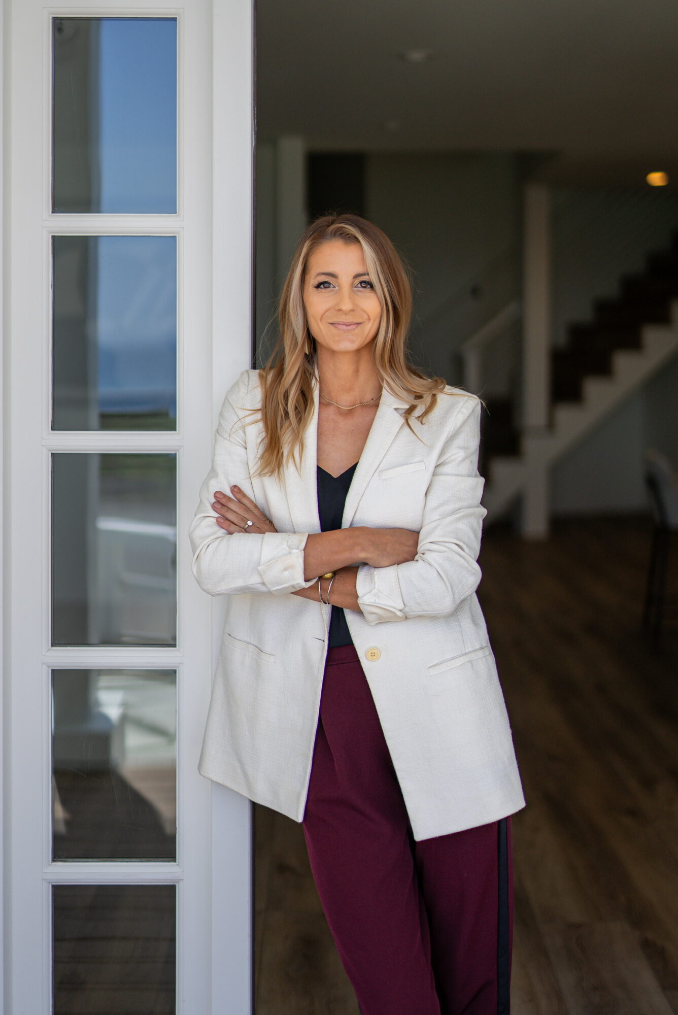 Woman with long hair in a white blazer and maroon pants standing indoors near a window, arms crossed, smiling.