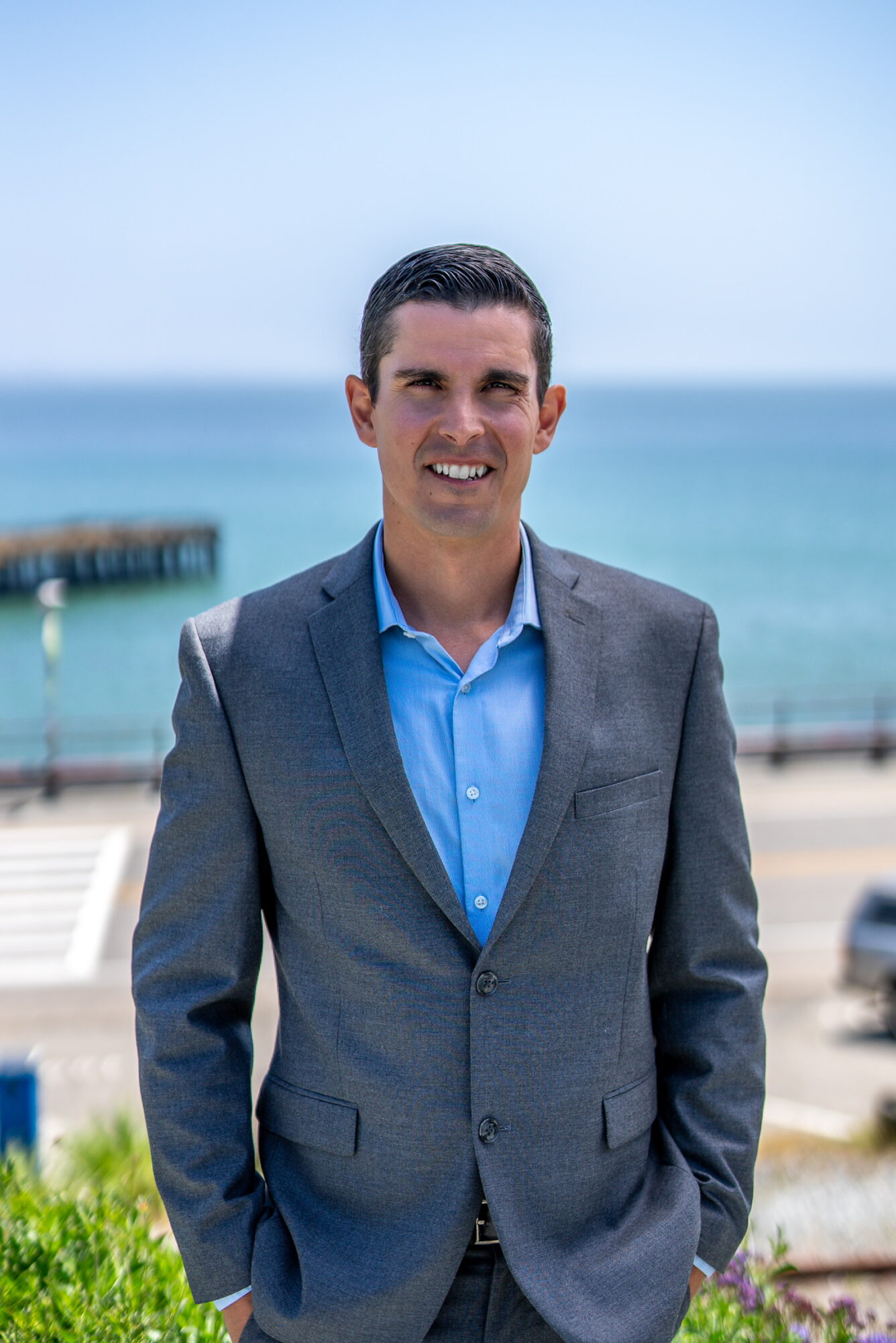 Man in a gray suit and blue shirt standing outdoors near the ocean with a pier in the background.