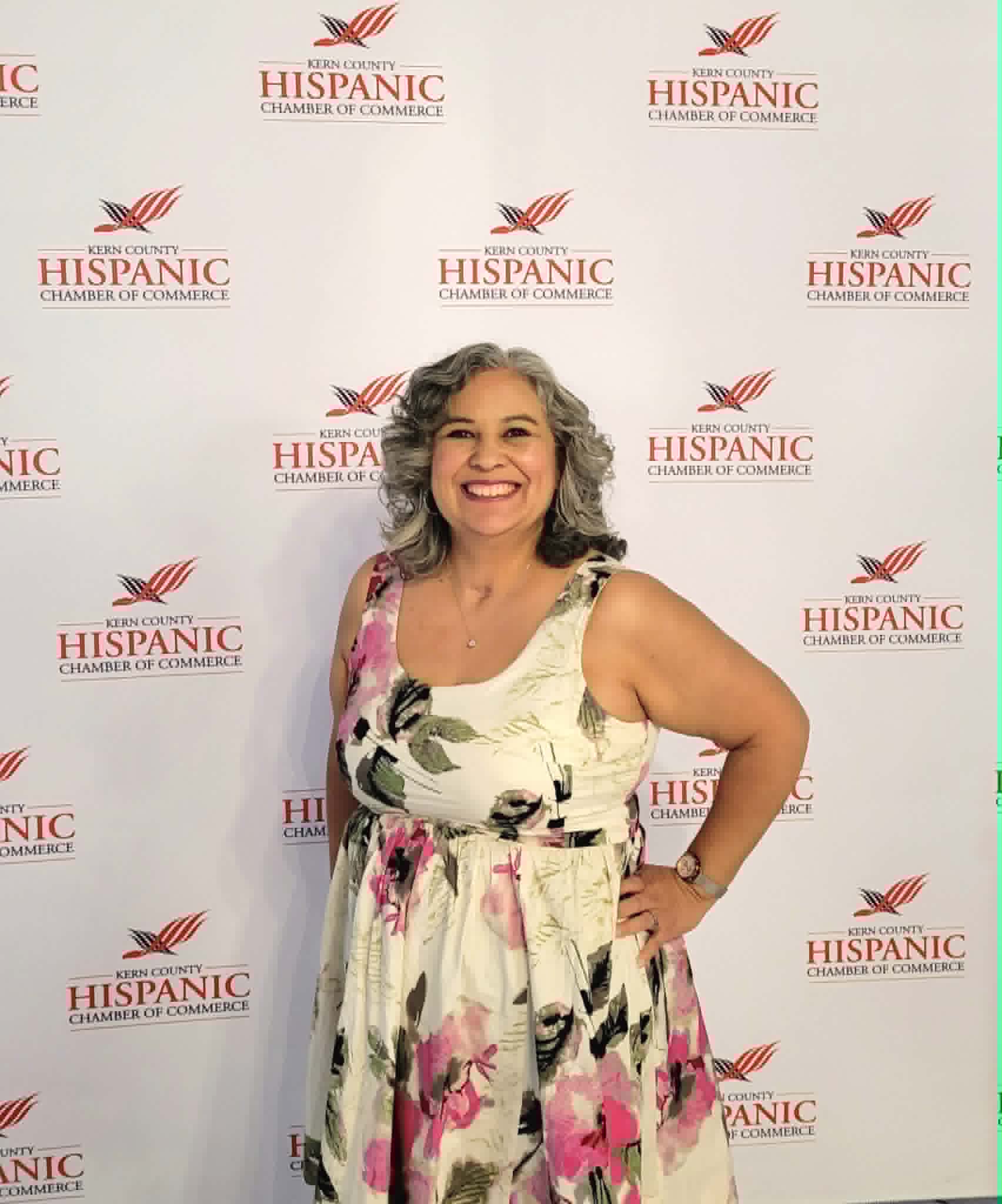 Smiling woman with curly gray hair in a floral dress standing in front of a Hispanic Chamber of Commerce backdrop.