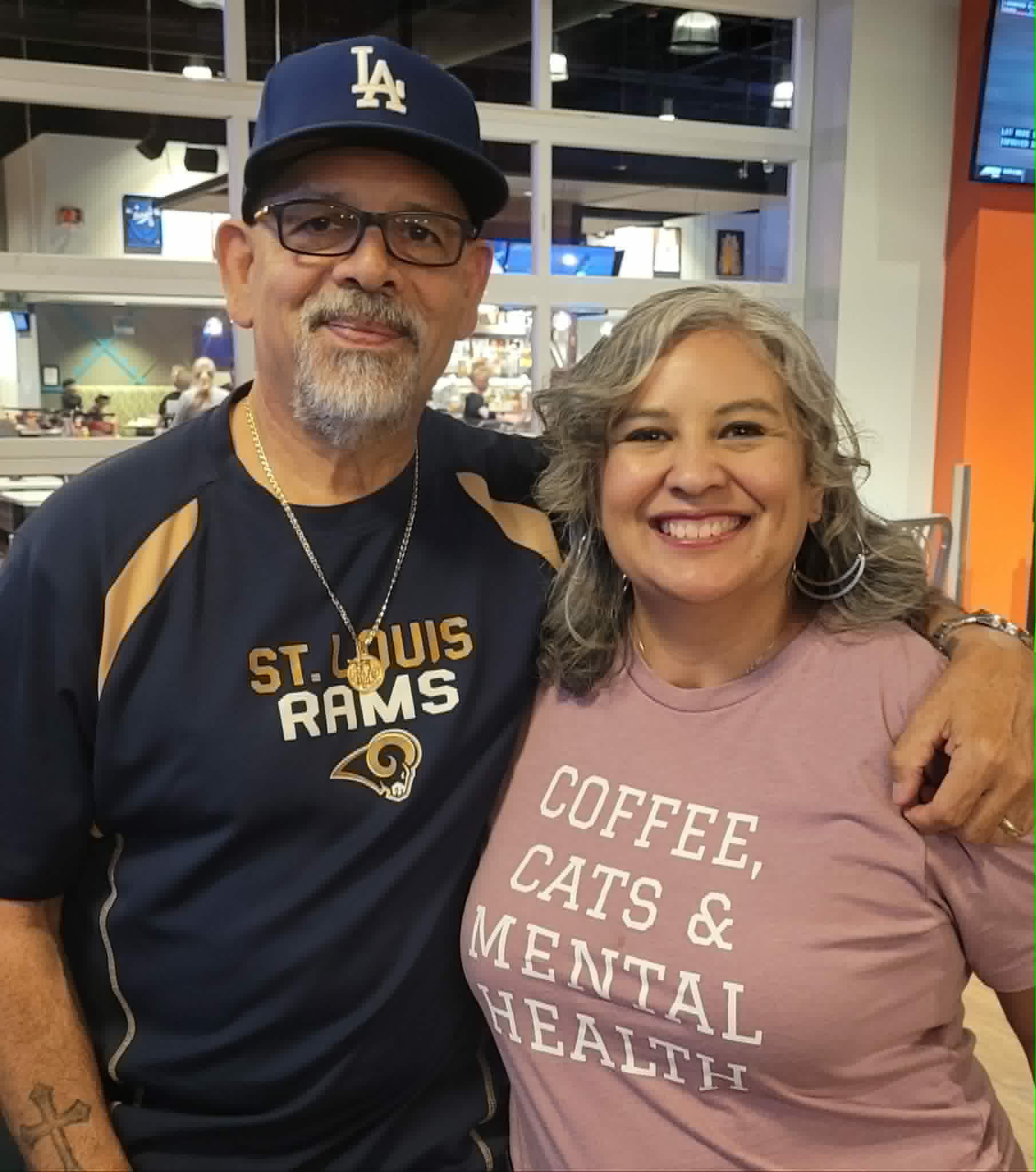 Two smiling people, one wearing a Los Angeles Dodgers cap and sports jersey, the other in a pink shirt, pose together indoors.