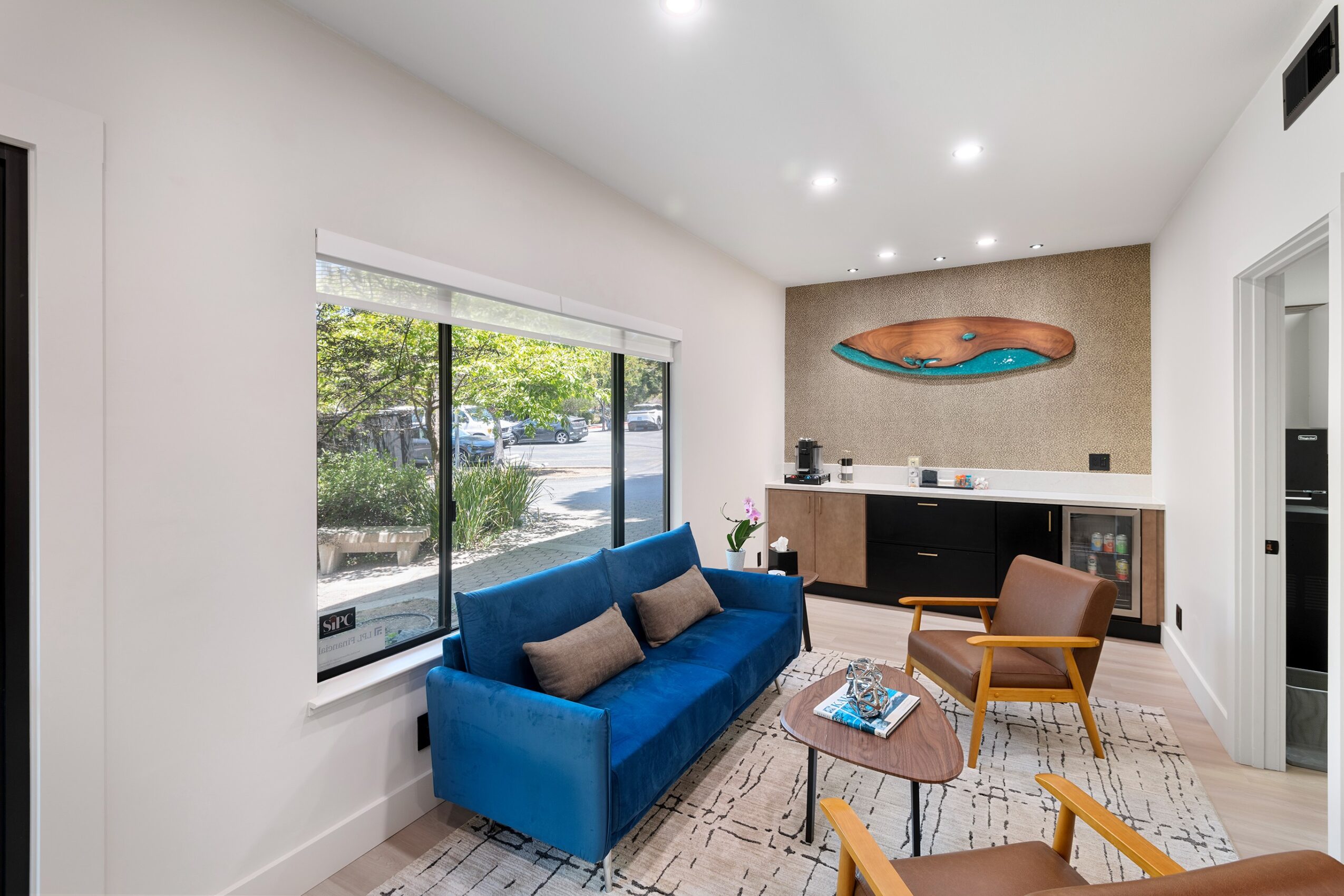 Living room with blue sofa, wooden chairs, window, and a wall-mounted art piece, bright and modern interior.