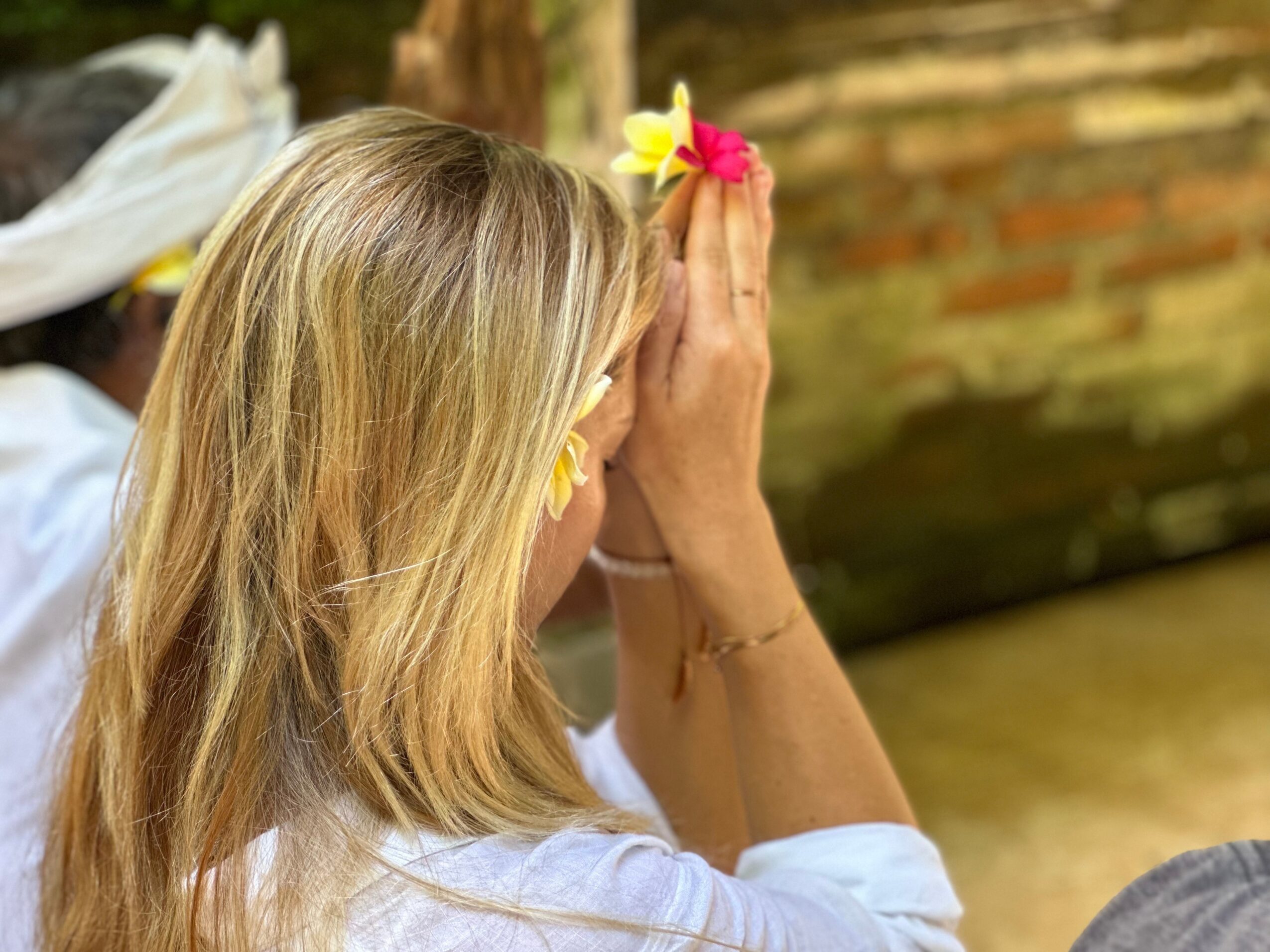 Woman with blonde hair holds flowers in a prayer position outdoors, with a brick wall in the background.