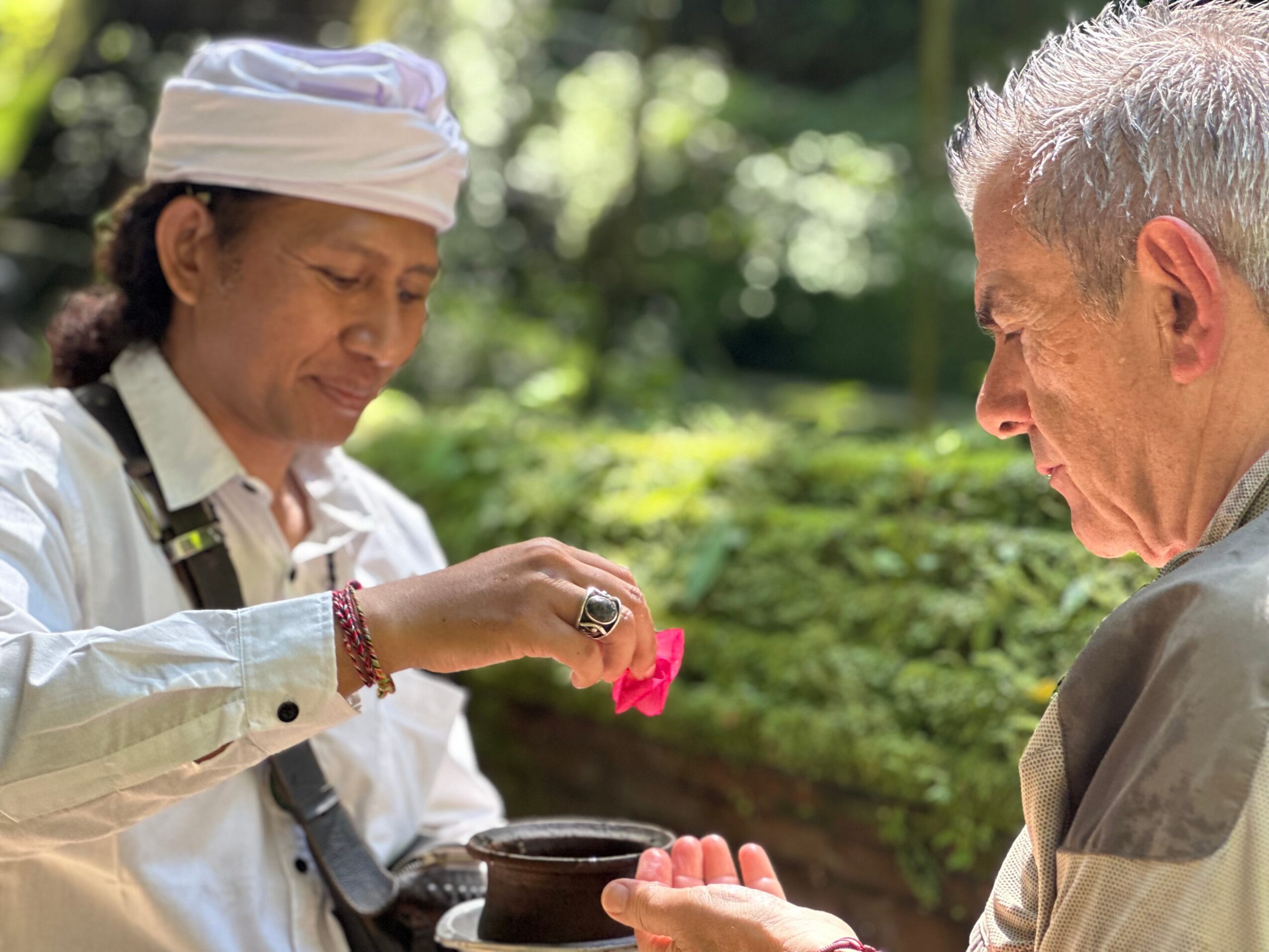Two people outdoors, one holding a pink object over a bowl, surrounded by greenery.