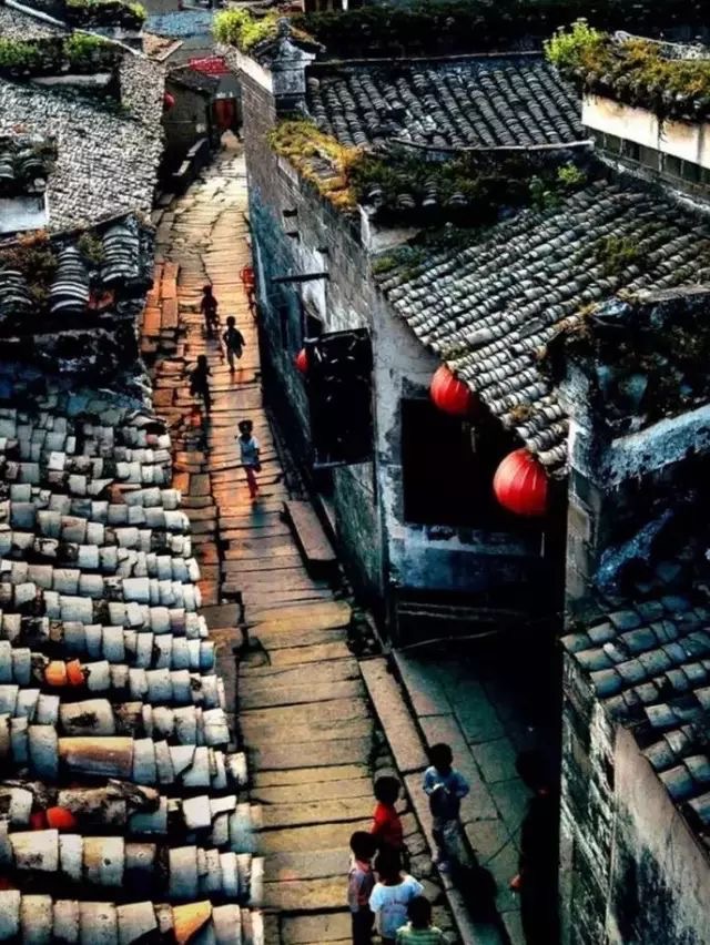 Narrow alleyway with traditional tiled roofs, people walking, and red lanterns hanging on a building wall.