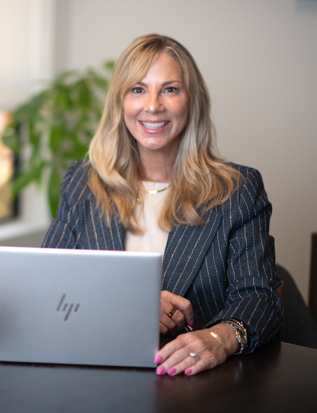 Woman with blonde hair smiling, sitting at desk with laptop, wearing a pinstripe blazer and white top.