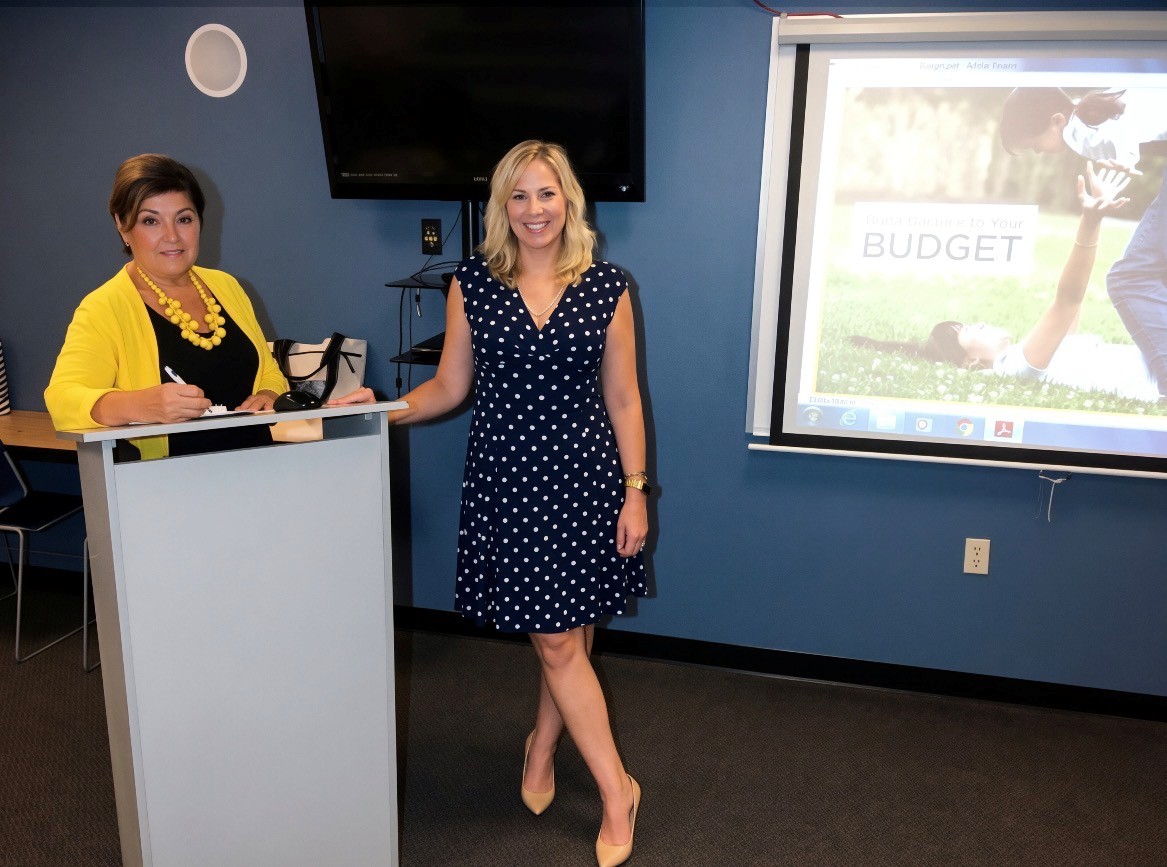 Two women standing in a room with a presentation screen, one at a podium, the other smiling in a polka dot dress.