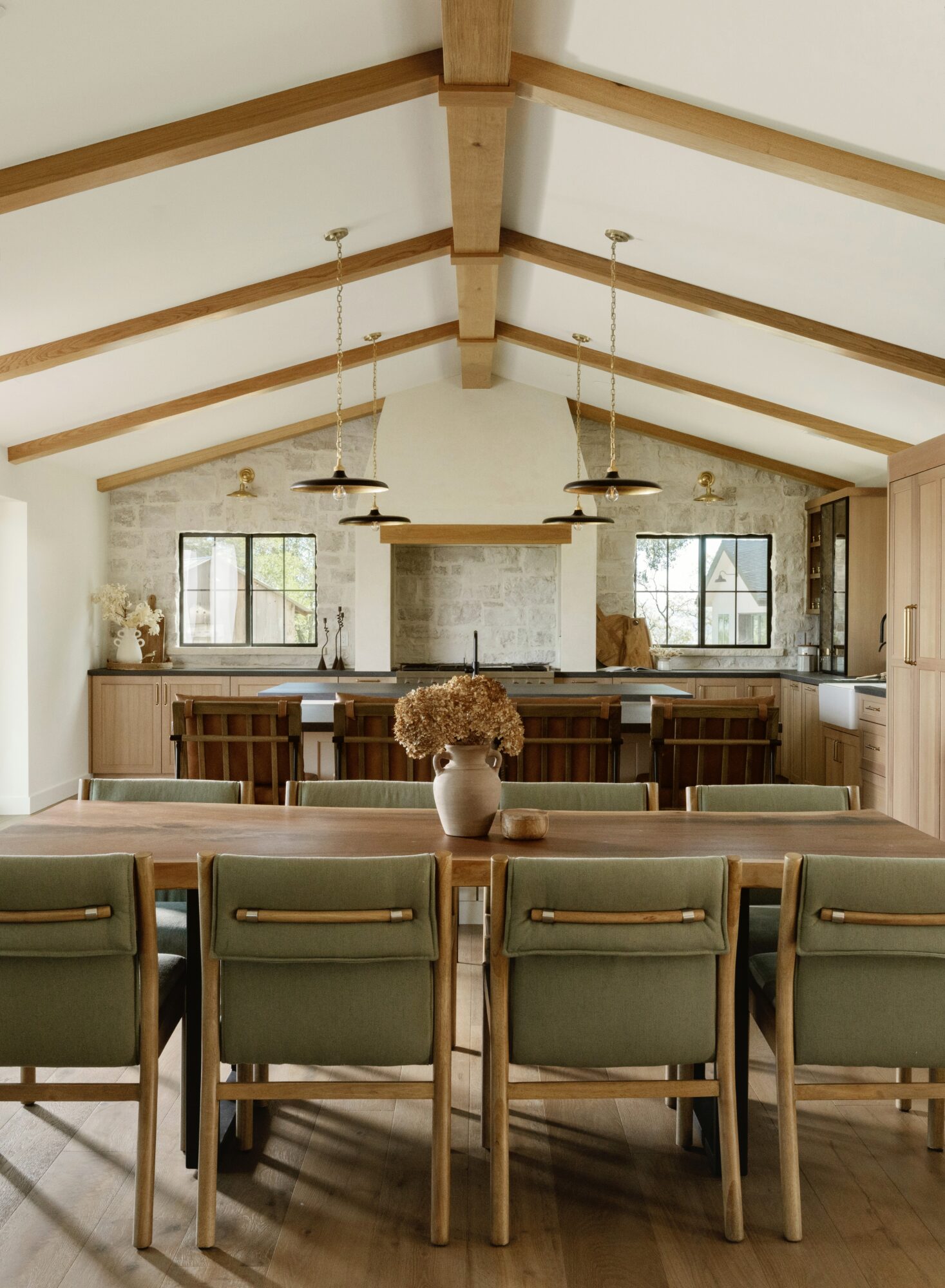 Interior of a dining area with a high, vaulted ceiling with exposed wooden beams, large windows, and a long wooden table with chairs.