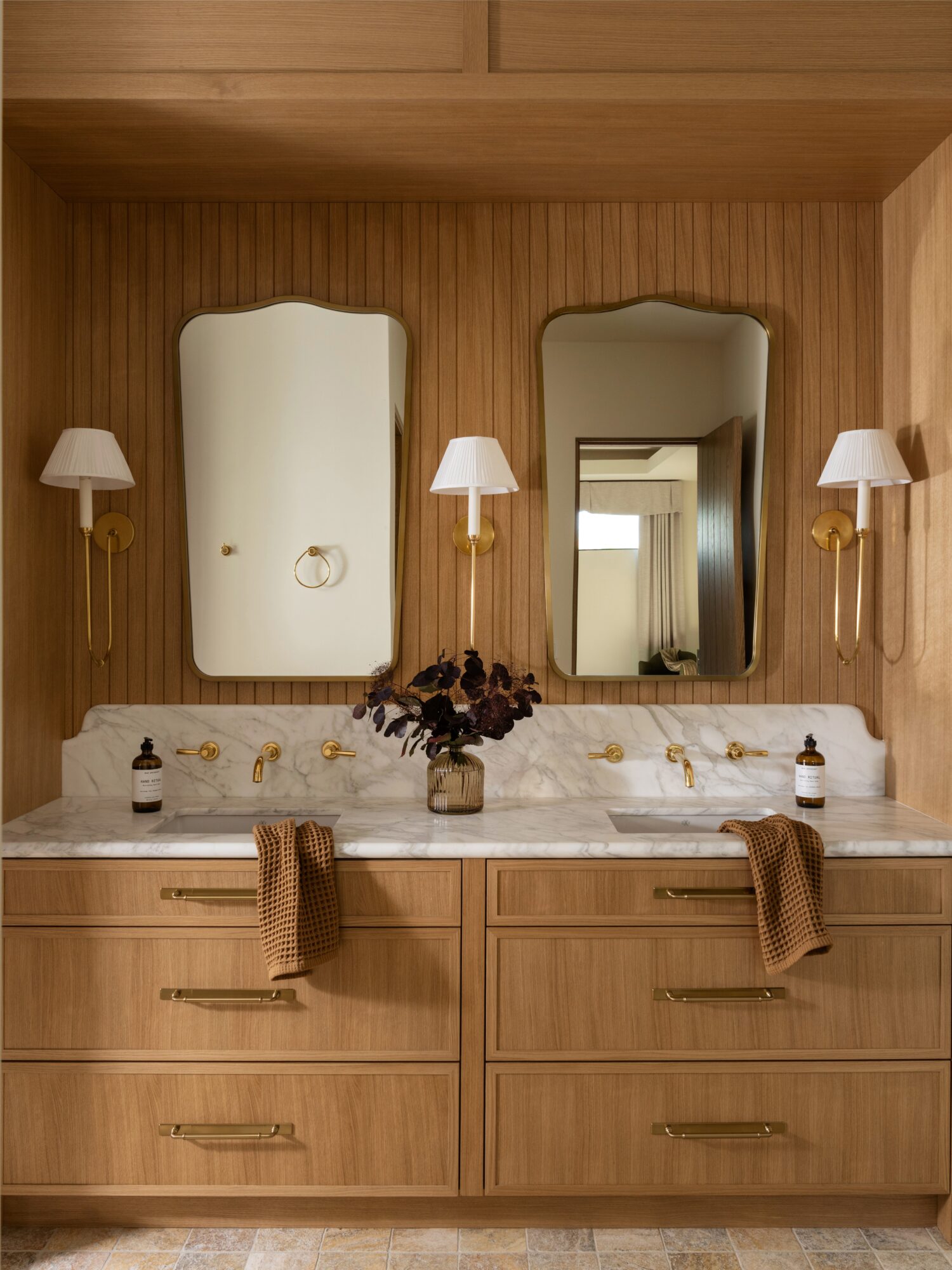 Wood-paneled bathroom with two mirrors, wall-mounted lights, and a marble countertop with towels and a flower arrangement.