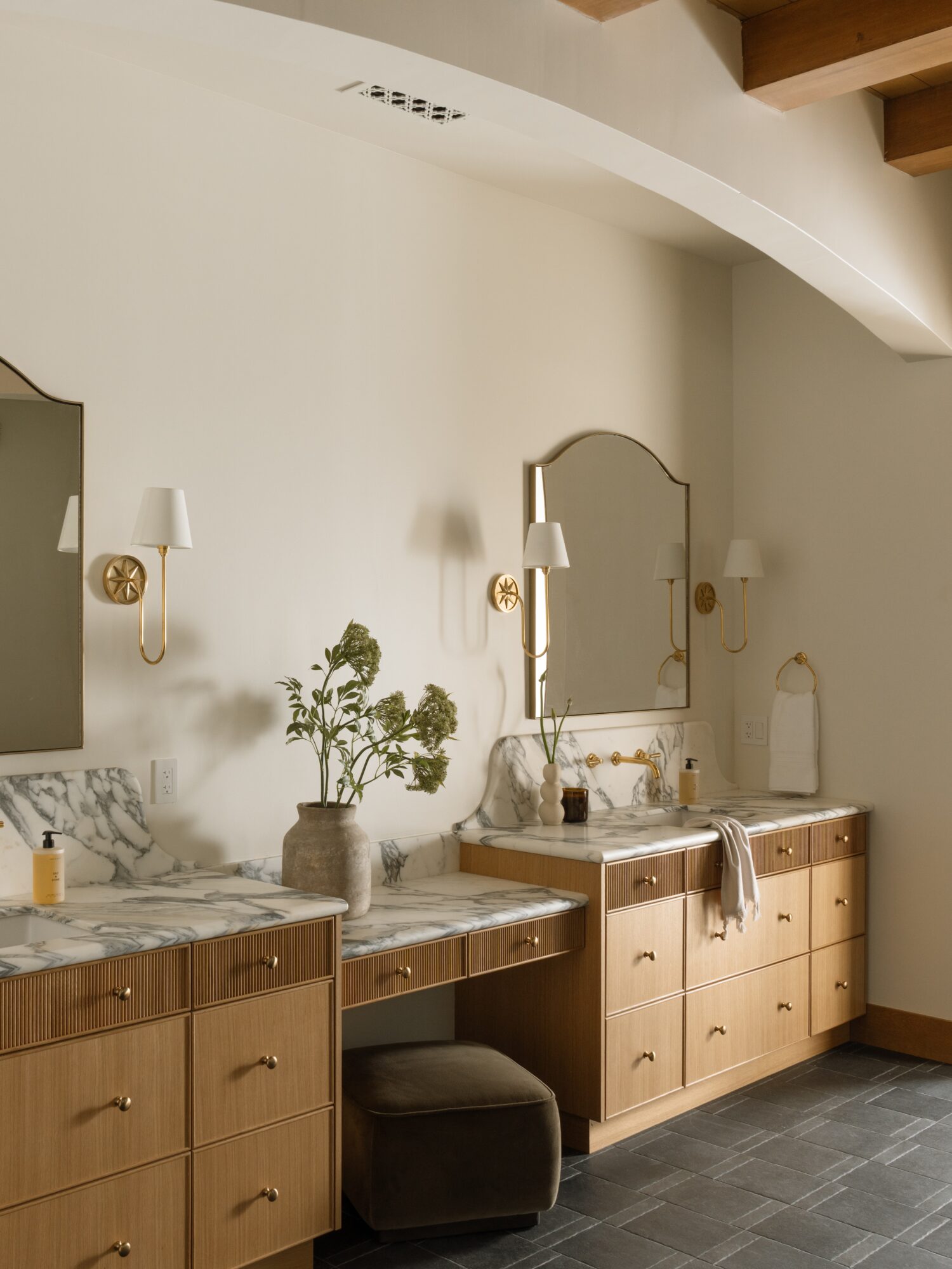 Bathroom vanity with mirror, plants, and towels, featuring wooden cabinets and a marble countertop, in a well-lit room.