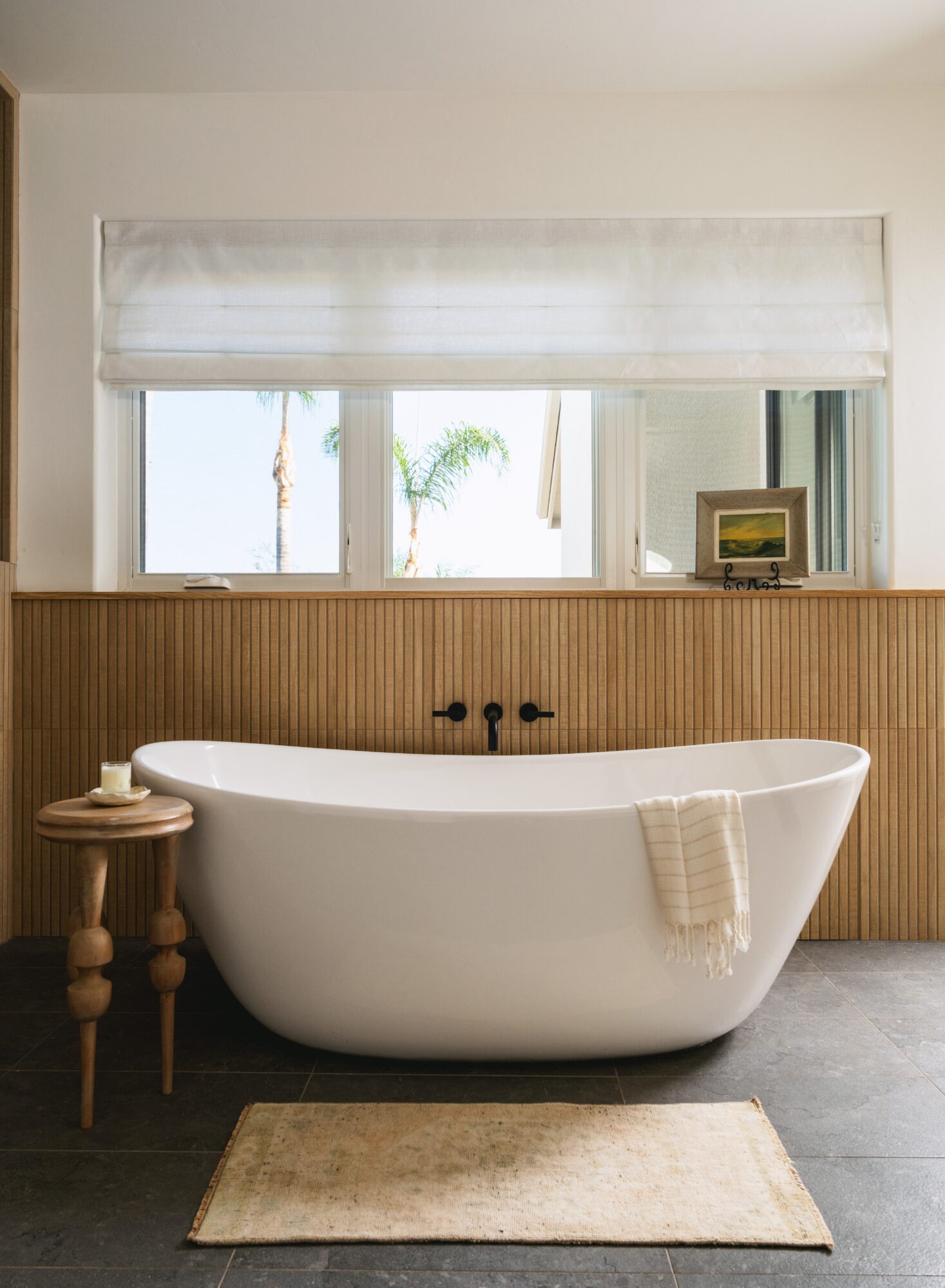 Freestanding white bathtub with a towel, wooden side table, window with blinds, and framed picture, in a bathroom setting.