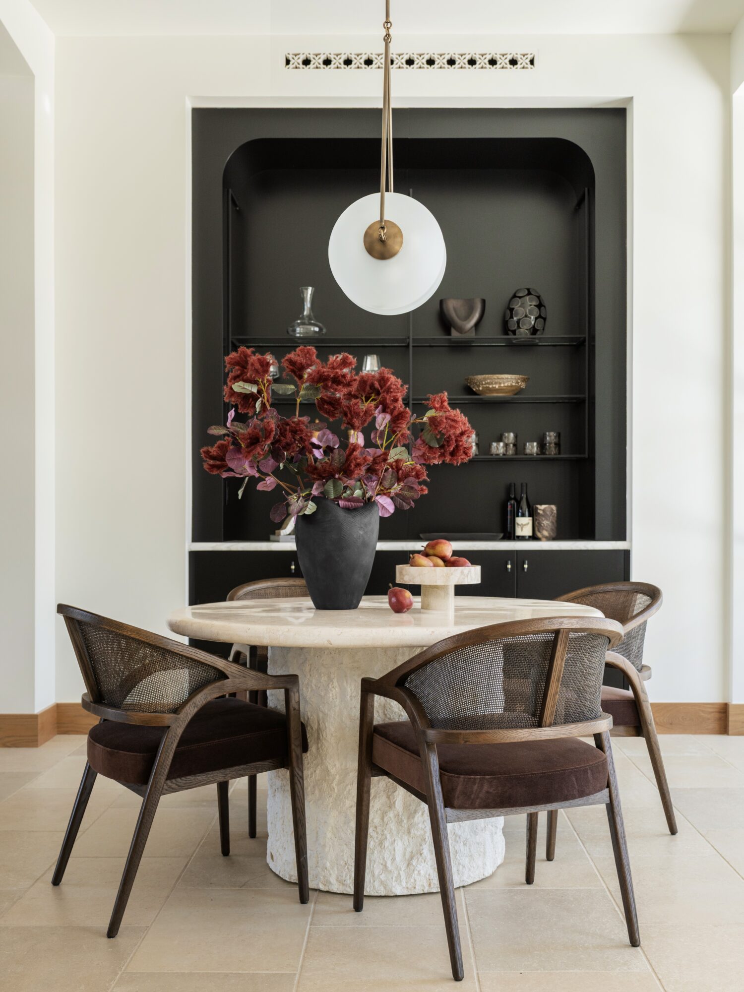 Round dining table with four chairs, a vase of red flowers, and a hanging light fixture, against a black built-in shelf.