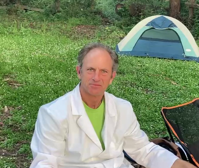 Man sitting outdoors near a tent, wearing a white jacket and green shirt, with trees and grass in background.
