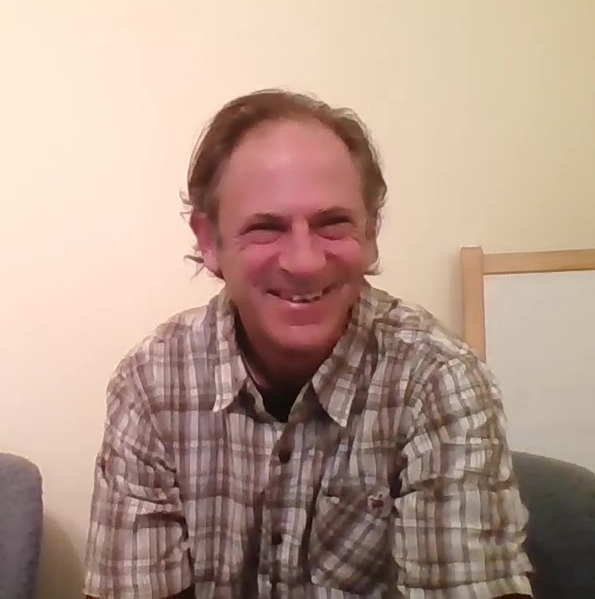 Smiling man with light brown hair wearing a plaid shirt, sitting indoors against a plain wall.