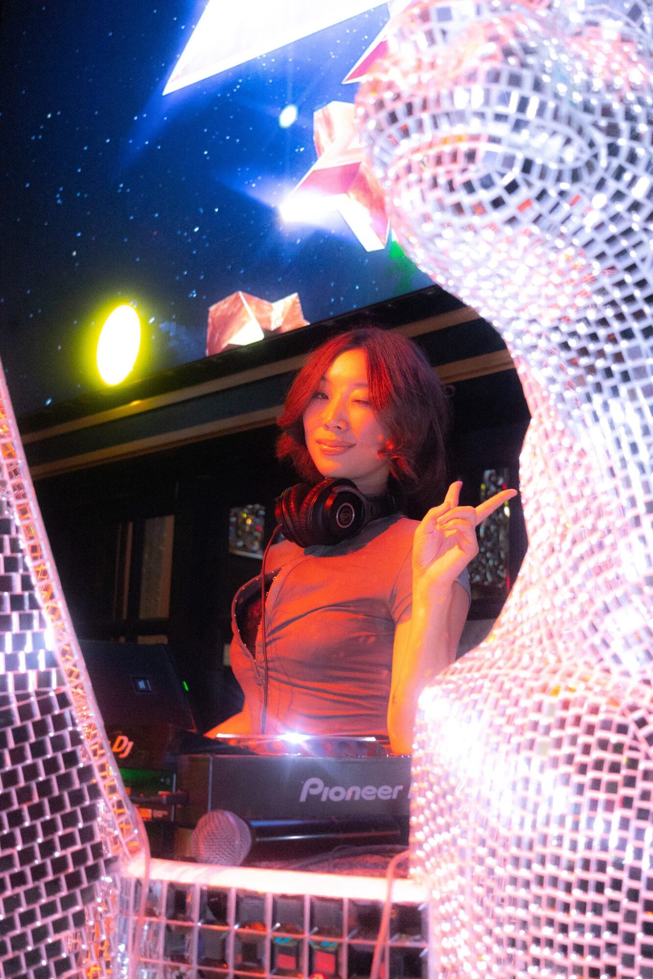 Female DJ with headphones smiling and making a peace sign at a colorful nightclub with disco decorations.