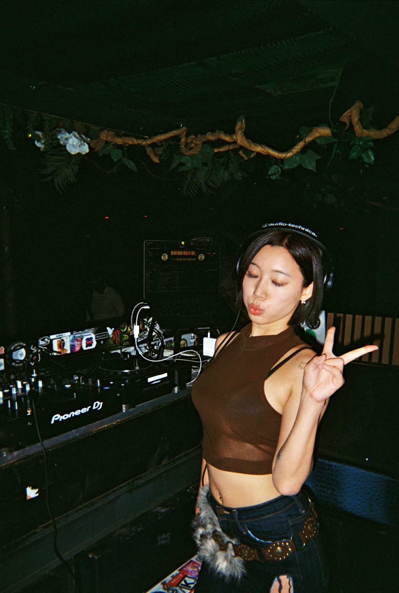 Young woman with headphones making a peace sign at a DJ setup in a dark environment.