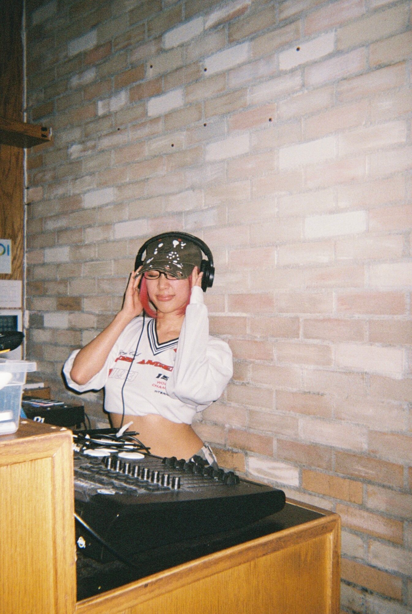 Person wearing headphones and sunglasses standing behind DJ equipment against a brick wall.