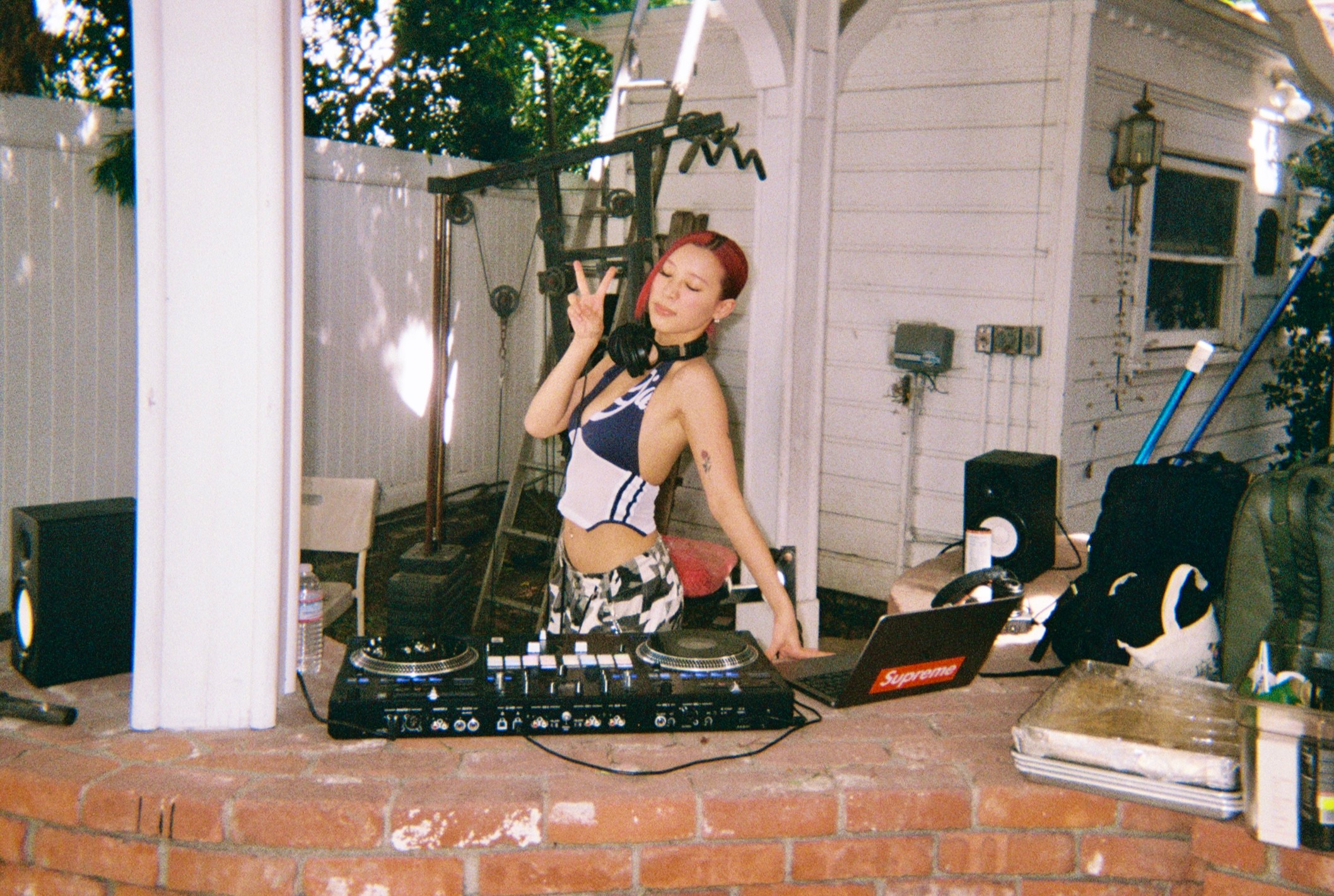 Person with red hair DJing outdoors, wearing a striped top, surrounded by equipment, in a backyard setting.