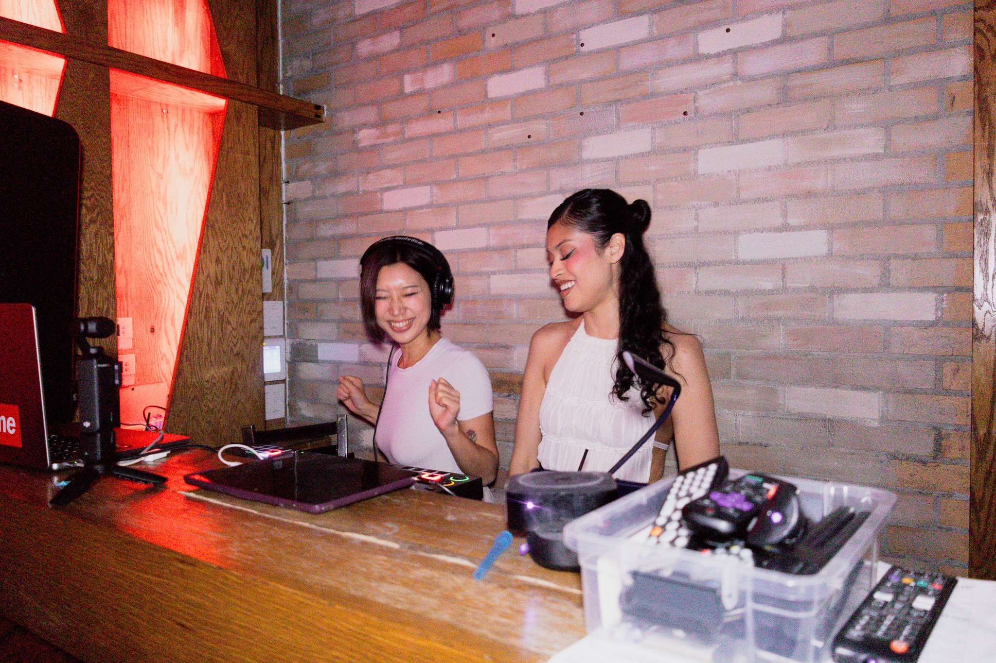 Two women are smiling and talking at a wooden bar counter with a brick wall background.
