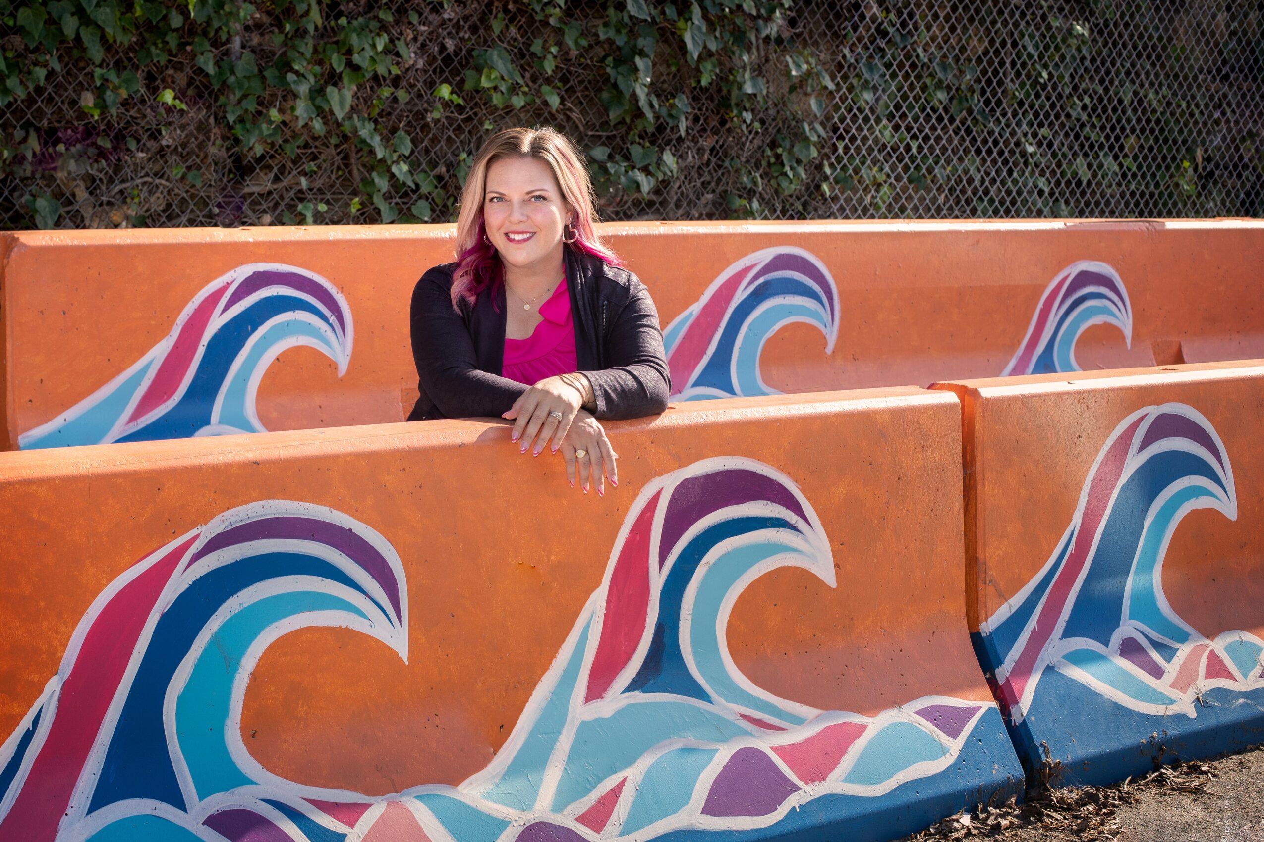 Woman with blonde hair smiling, sitting behind a colorful wave-themed barrier outdoors, with trees and a fence in background.