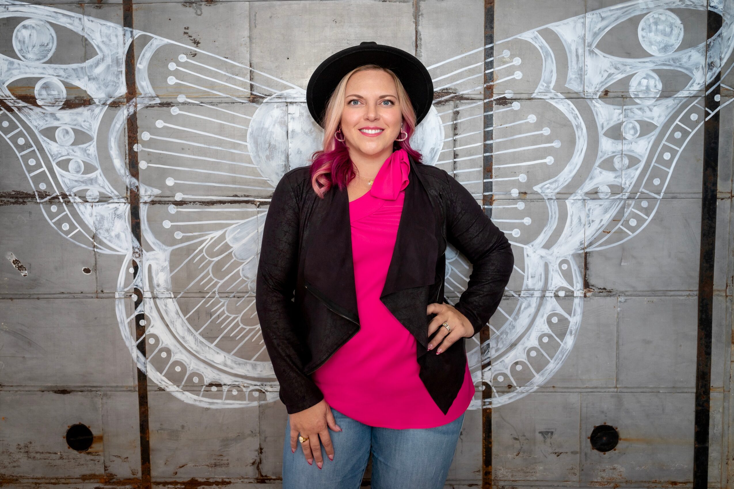 Woman with pink hair, black hat, pink top, black jacket, standing in front of butterfly mural, smiling, hands on hips.