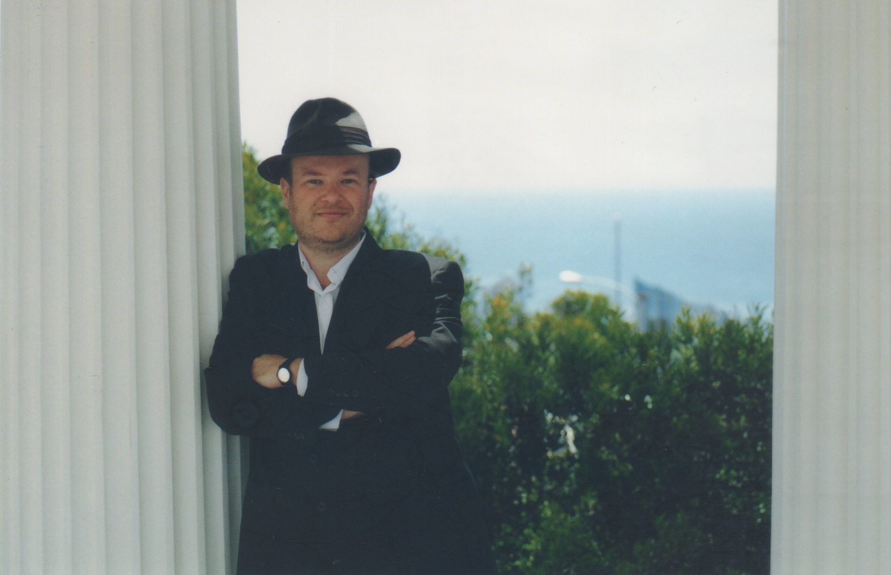 Man in a suit and hat standing outdoors with a view of the ocean and greenery behind him.
