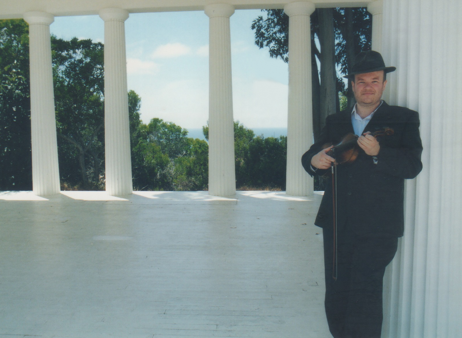 Person in black suit and hat holding a guitar, standing on a white porch with columns and trees in background.