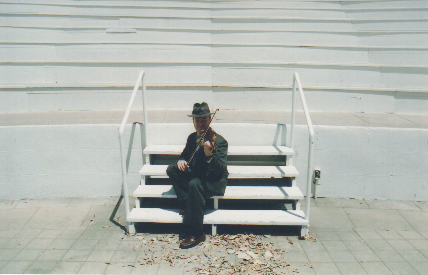 Person sitting on stairs, holding a violin, outdoors, with white stairs and railing behind them.