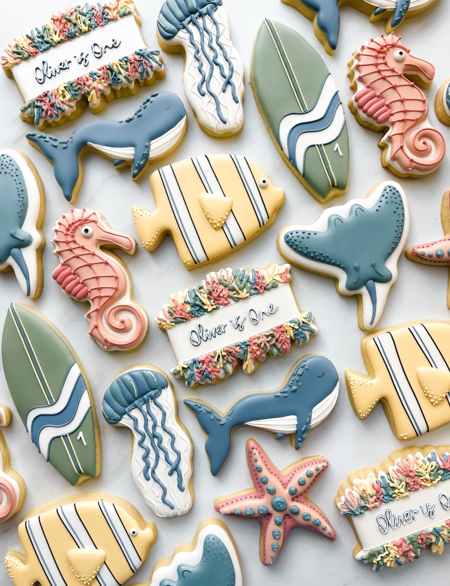 Assorted ocean-themed cookies with whales, seahorses, starfish, surfboards, and floral banners, arranged on a white background.