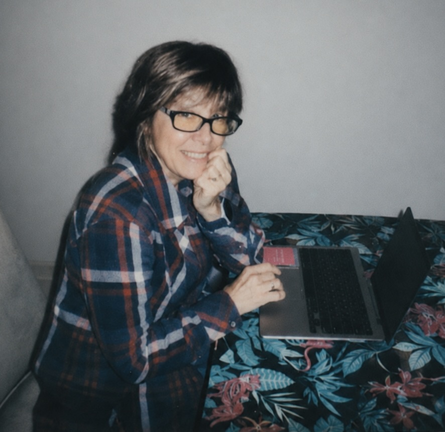 Woman with glasses smiling at laptop, sitting at a table with a floral tablecloth, hand near chin, indoors.