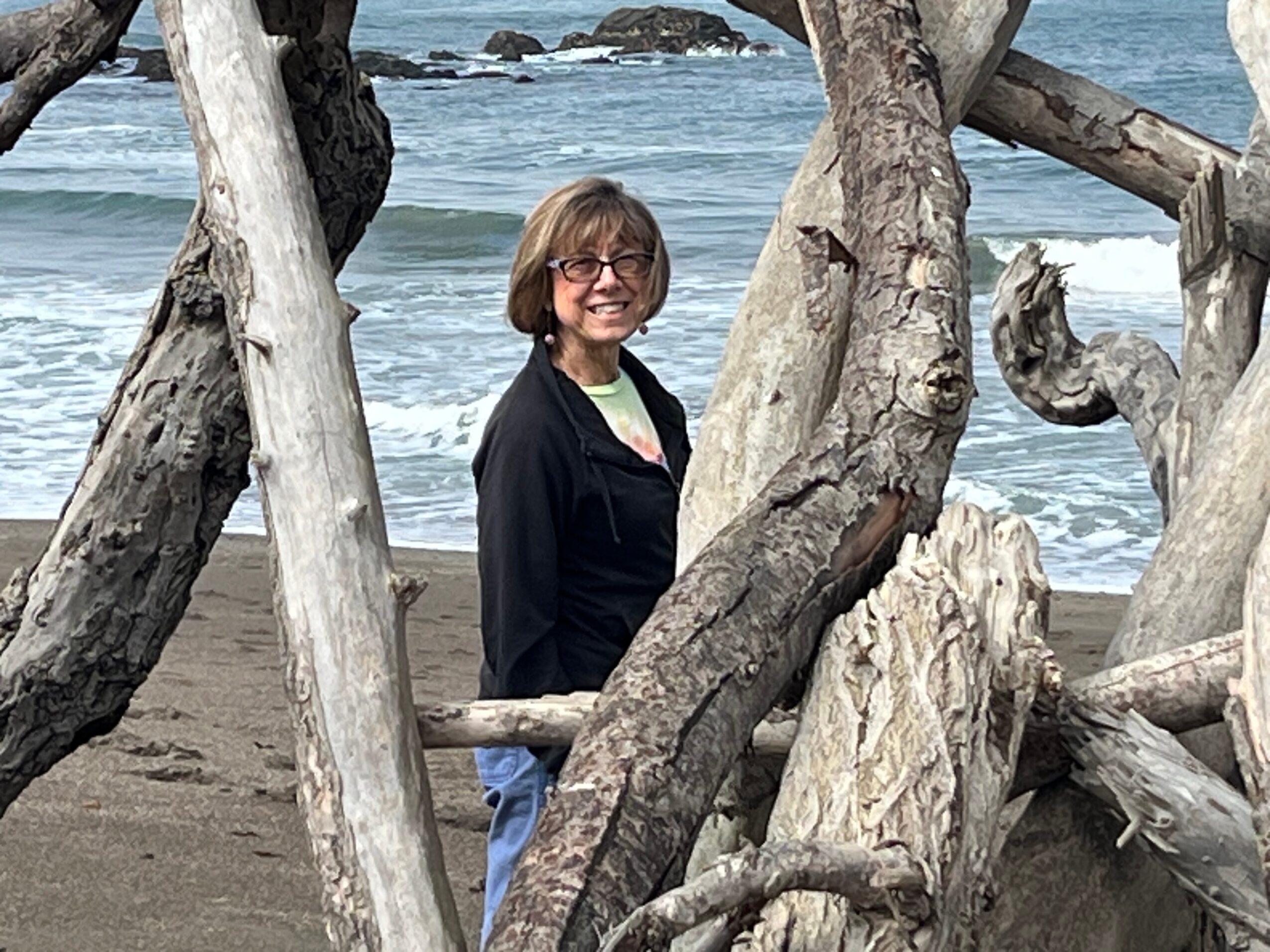 Woman with glasses smiling on a beach, framed by large driftwood pieces, ocean waves in background.