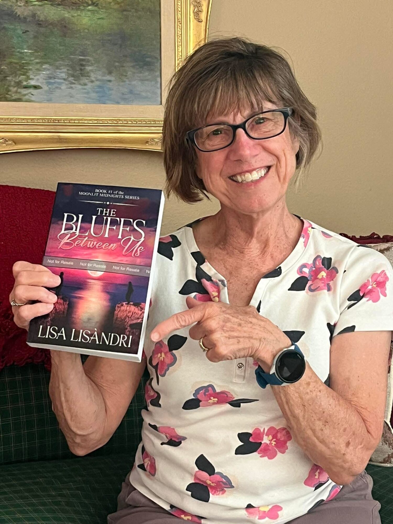 Smiling woman with glasses holding a book titled 'The Buffs' in a room with a painting and beige wall.