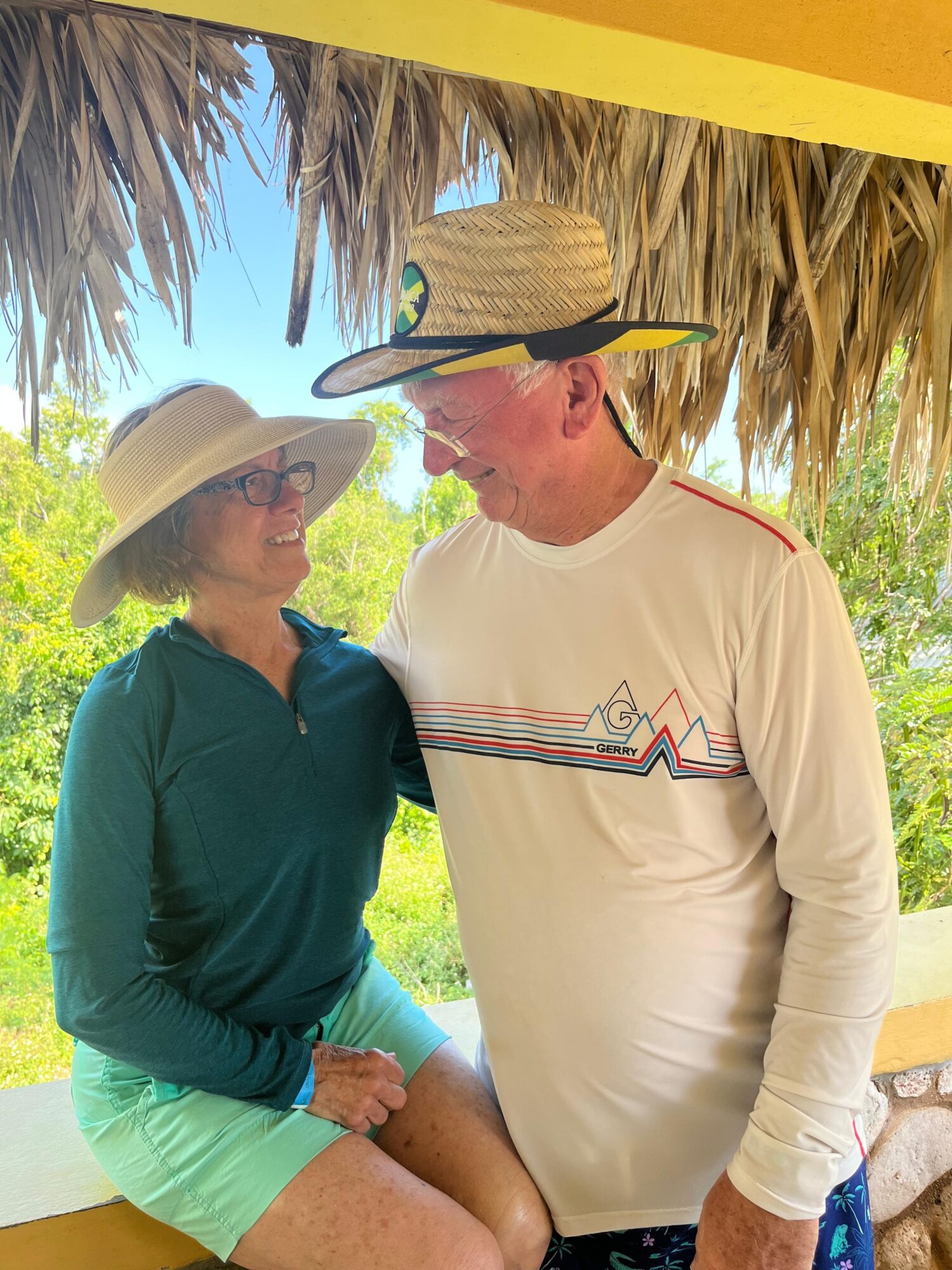 Two people wearing hats and sunglasses under a thatched roof, smiling at each other outdoors with trees and blue sky in background.