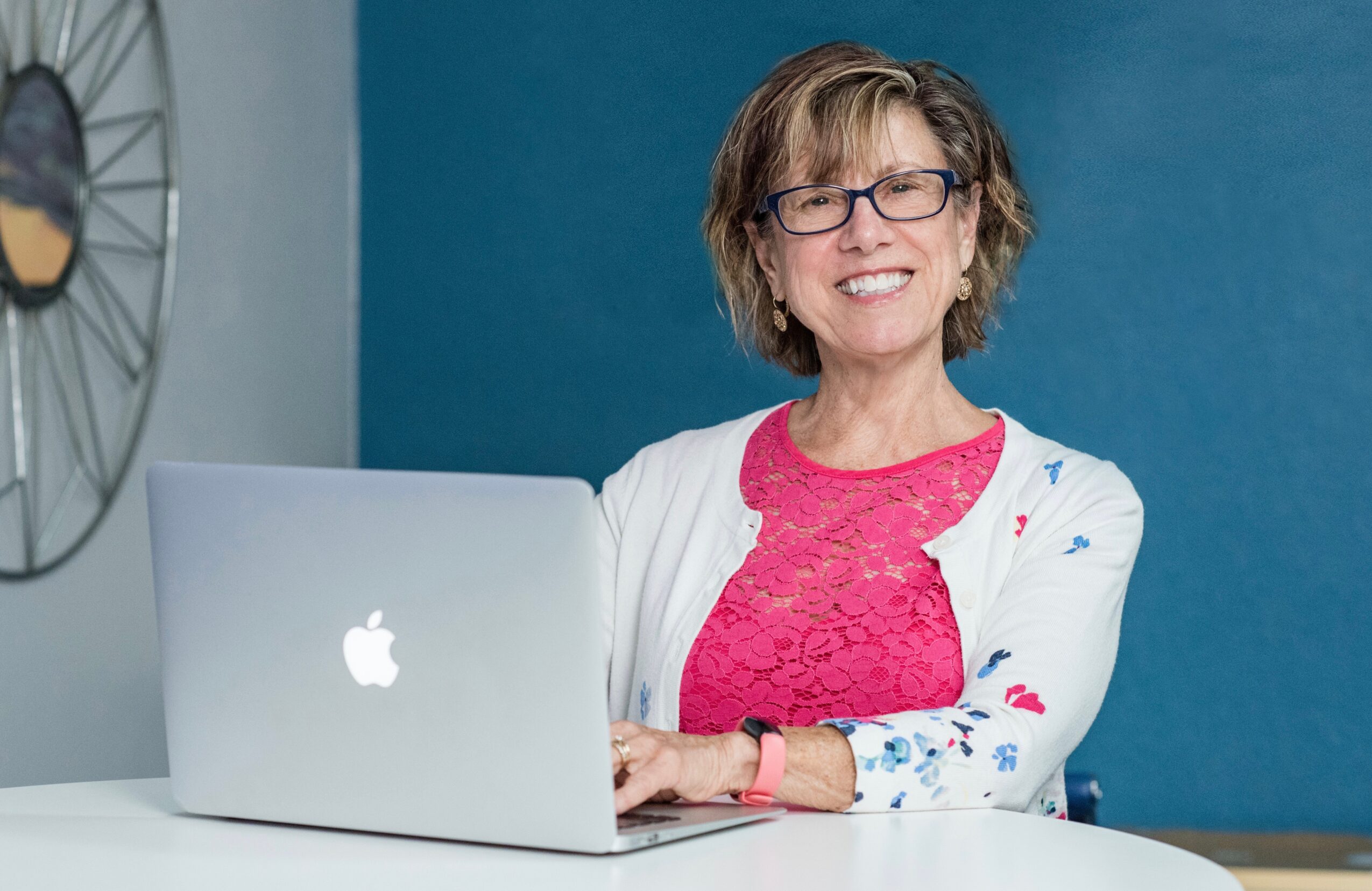 Smiling woman with glasses sitting at a table with a laptop, blue background, wearing a pink top and white cardigan.