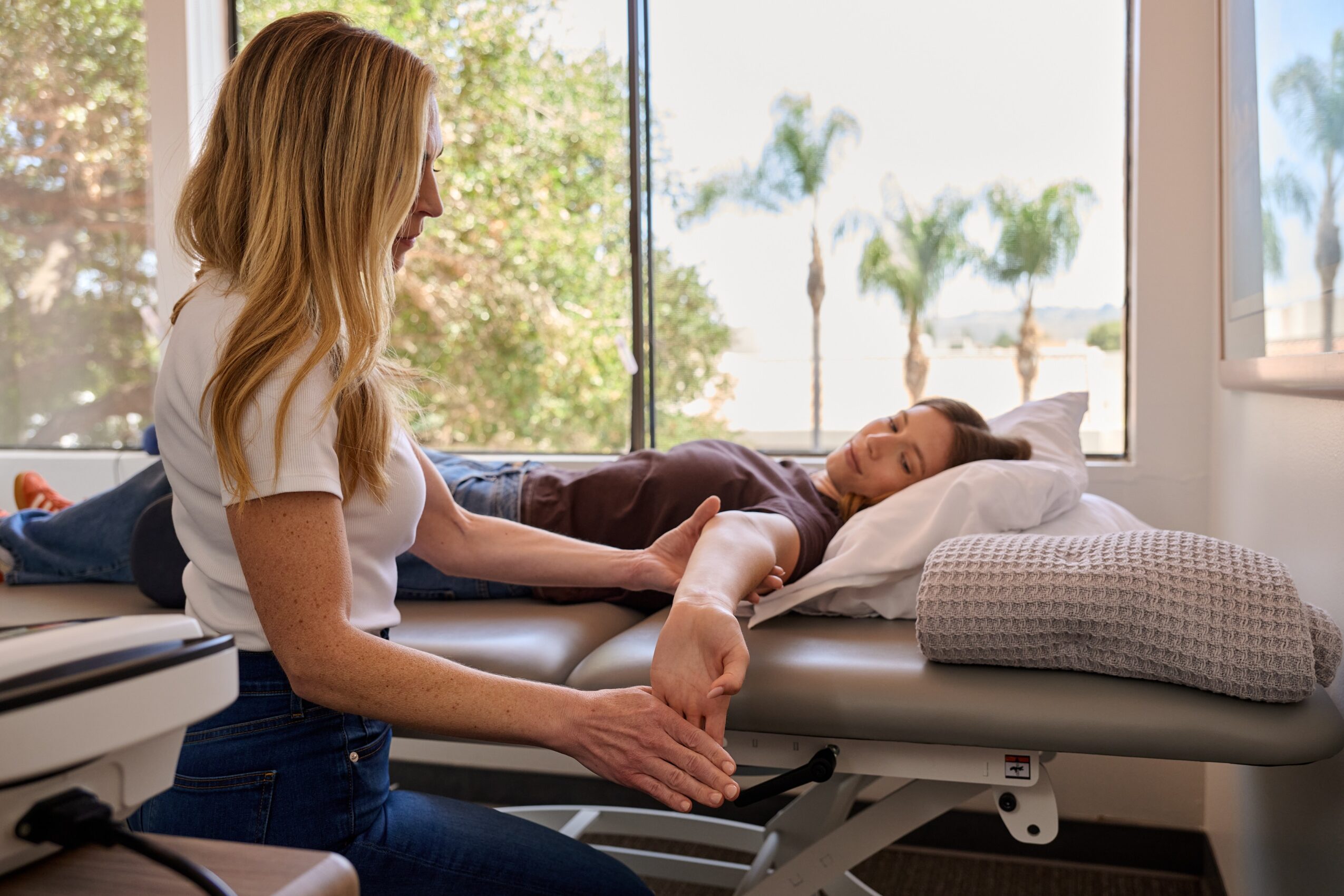 A woman holding a child's hand while the child lies on a medical examination table, with large windows and palm trees outside.