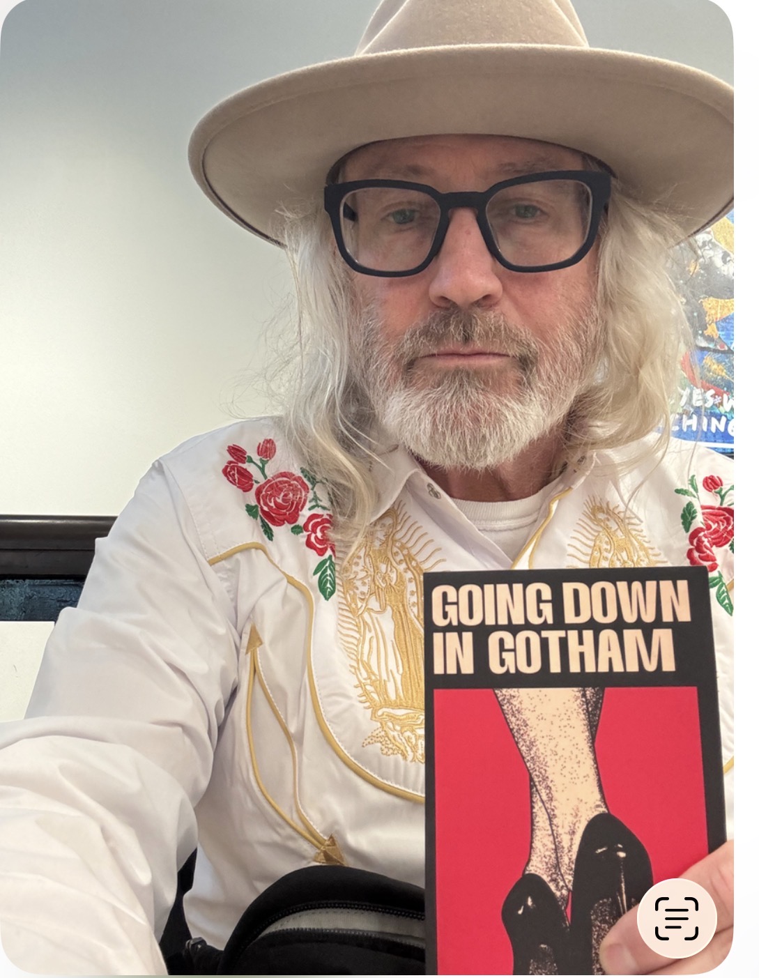 Older man with long gray hair, beard, glasses, wearing a wide-brimmed hat and embroidered white shirt, holding a book titled 'Going Down in Gotham'.
