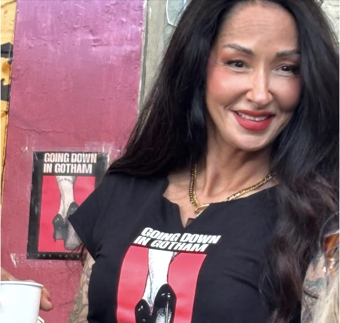 Woman with long dark hair smiling, wearing a black shirt with a graphic, standing outdoors near a pink wall.