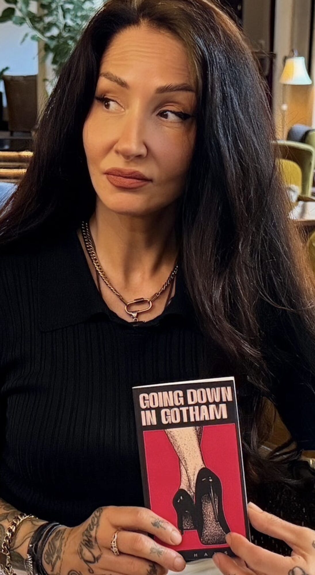 Woman with long dark hair holding a book titled 'Going Down in Gotham' in a room with warm lighting.