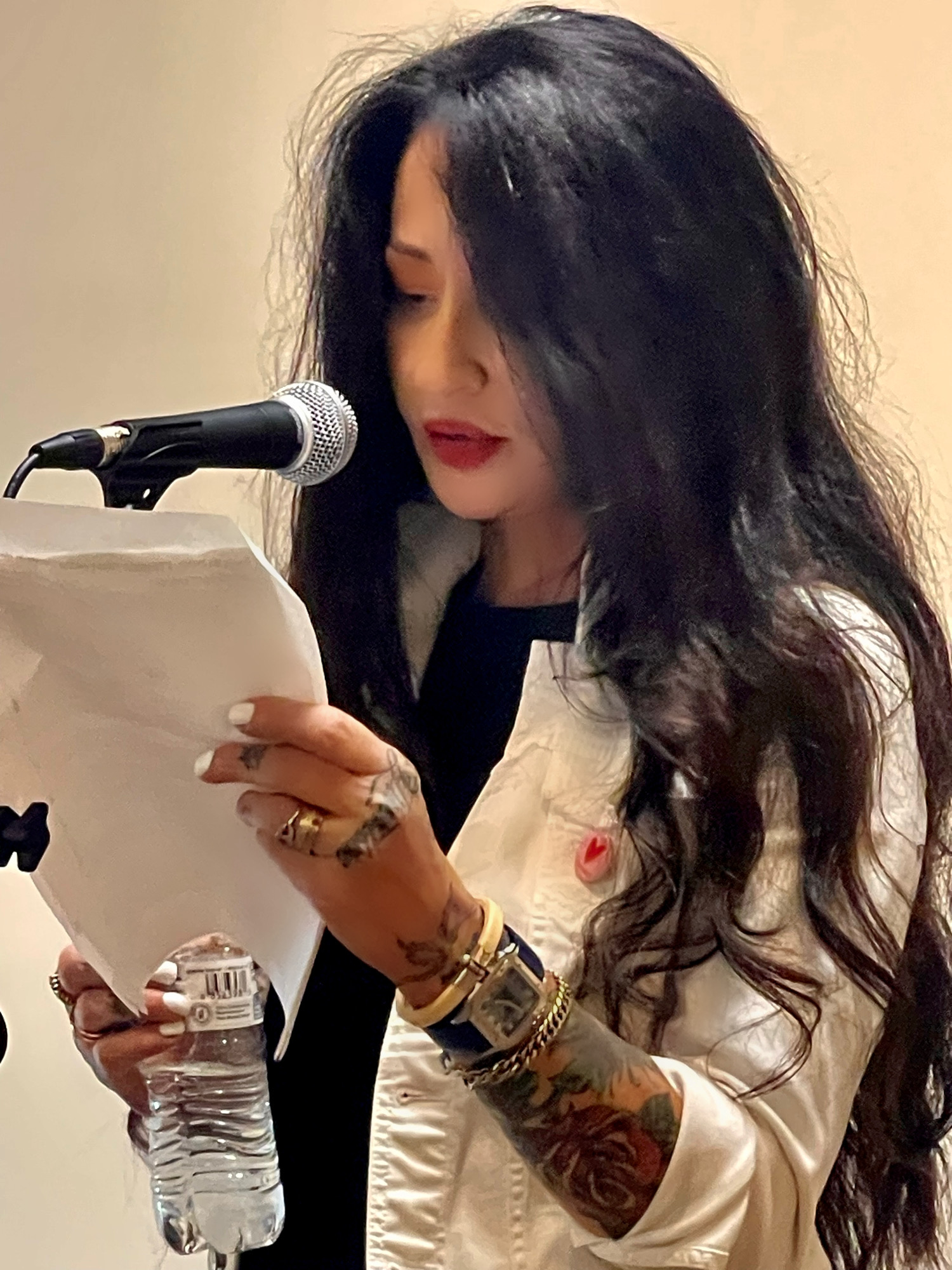 Woman with long dark hair reading from paper at microphone, holding water bottle, wearing white jacket, with tattoos on arm.